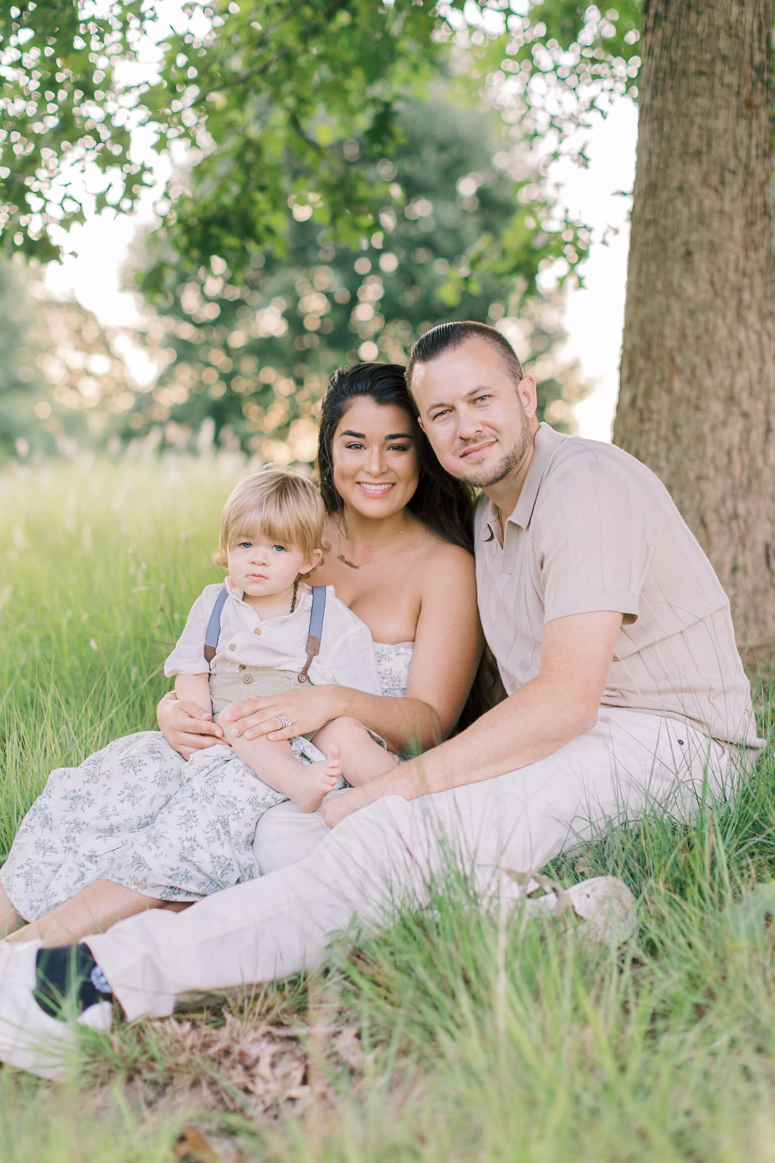 A mom and dad sit under a tree with their infant in their lap with big smiles at sunset after meeting pediatricians in raleigh