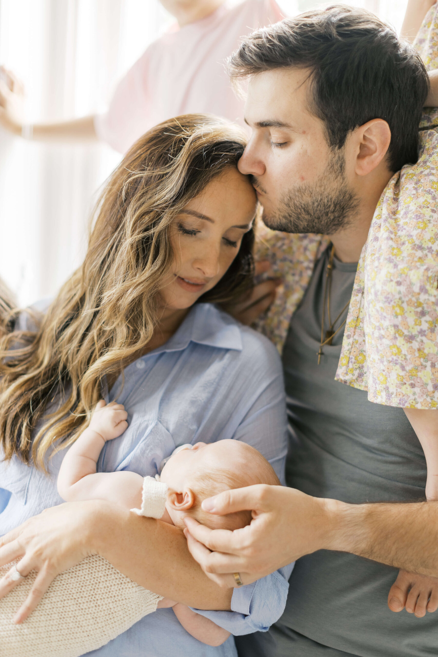A mom holding her newborn baby in her arms is kissed by dad as she leans into his chest under a window