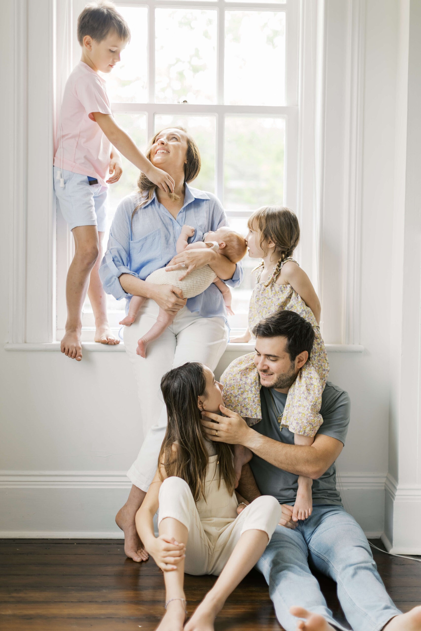 A mom and dad sit under a window as their three young children climb on them while holding a newborn thanks to a postpartum doulas in raleigh