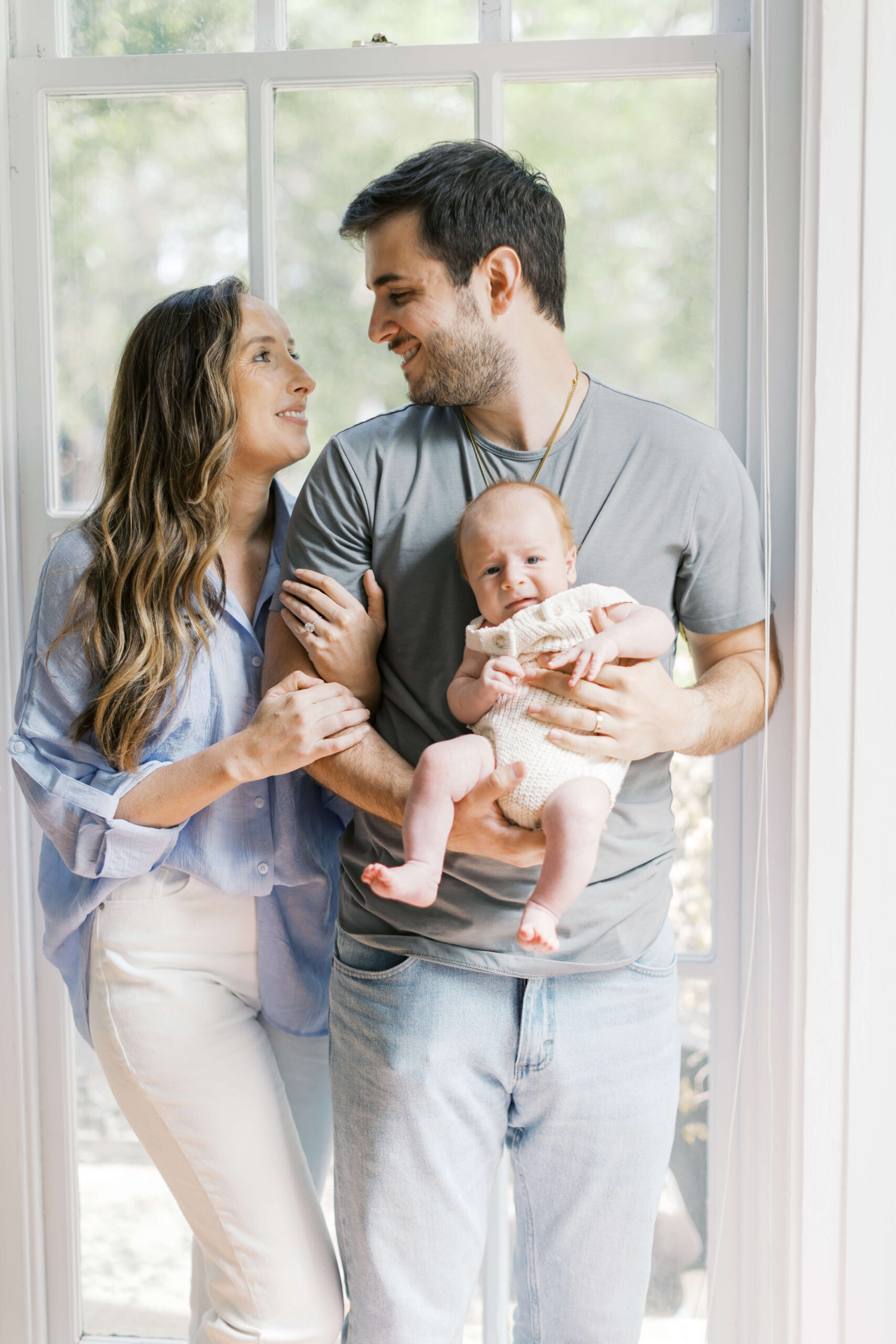 Happy new parents cuddle and lean in a window holding their newborn baby and smiling at each other thanks to postpartum doulas in raleigh