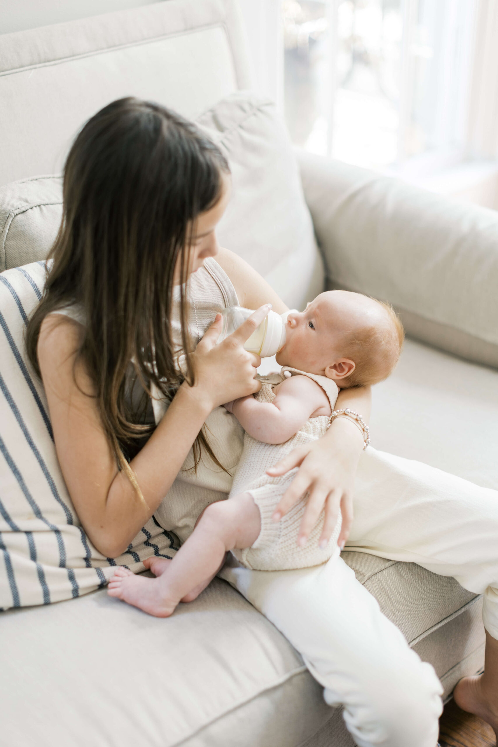 A young girl feeds her newborn baby sibling from a bottle while sitting on a couch after learning from postpartum doulas in raleigh