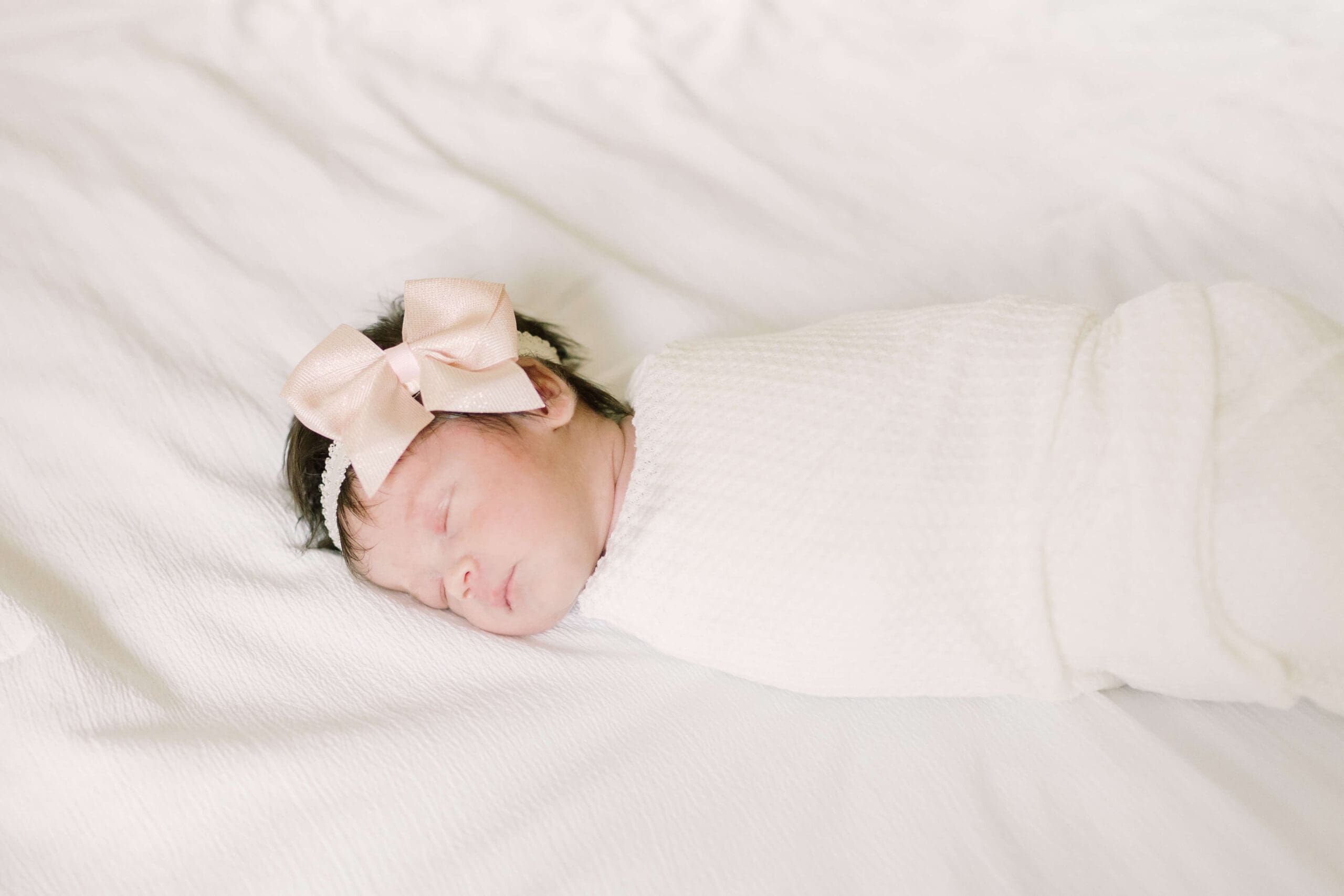 a newborn baby sleeps while swaddled in a cream cloth and wears a cream headband