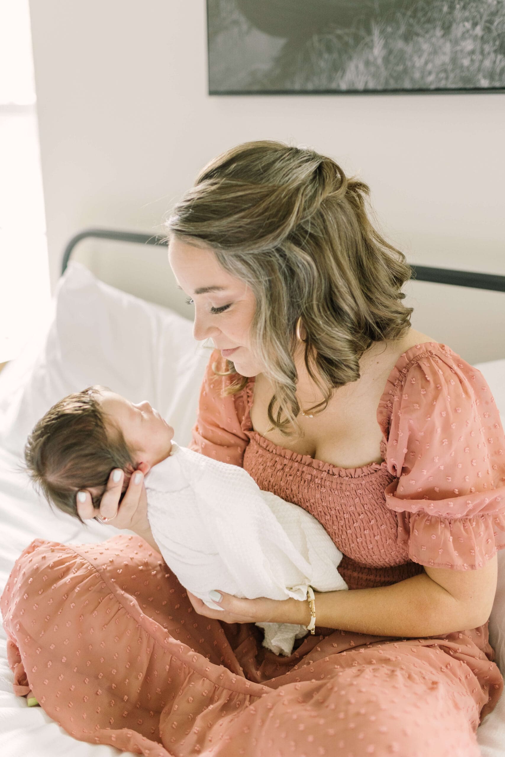 a new mom in a rust colored dress sits on a bed holding her newborn baby girl, born with the help of raleigh doulas