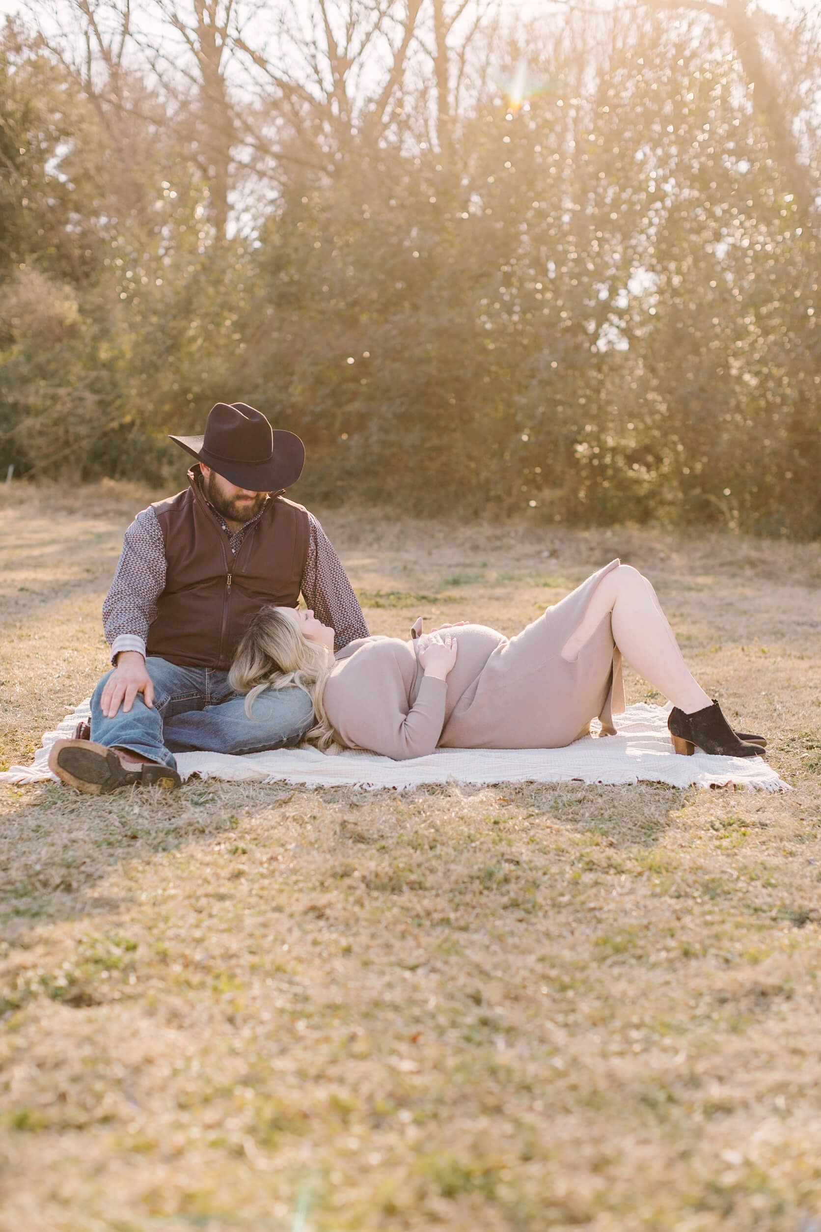 a mom to be rests her head in the lap of her husband in jeans and cowboy hat in a park at sunset