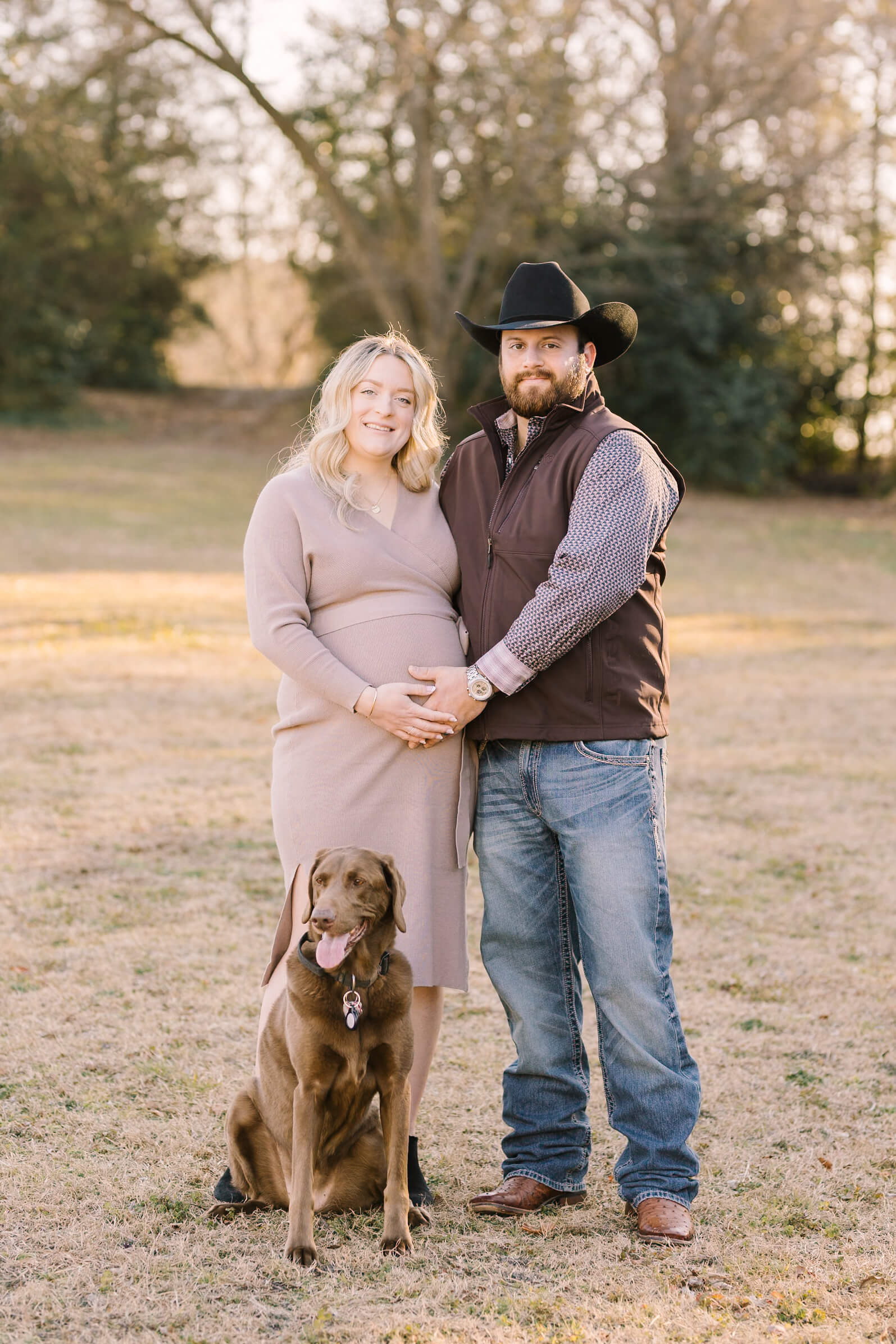 a mom to be in a brown dress stands in a field at sunset holding the bump with her husband in a vest and jeans with their brown dog sitting in front of them before their 3d ultrasound in raleigh