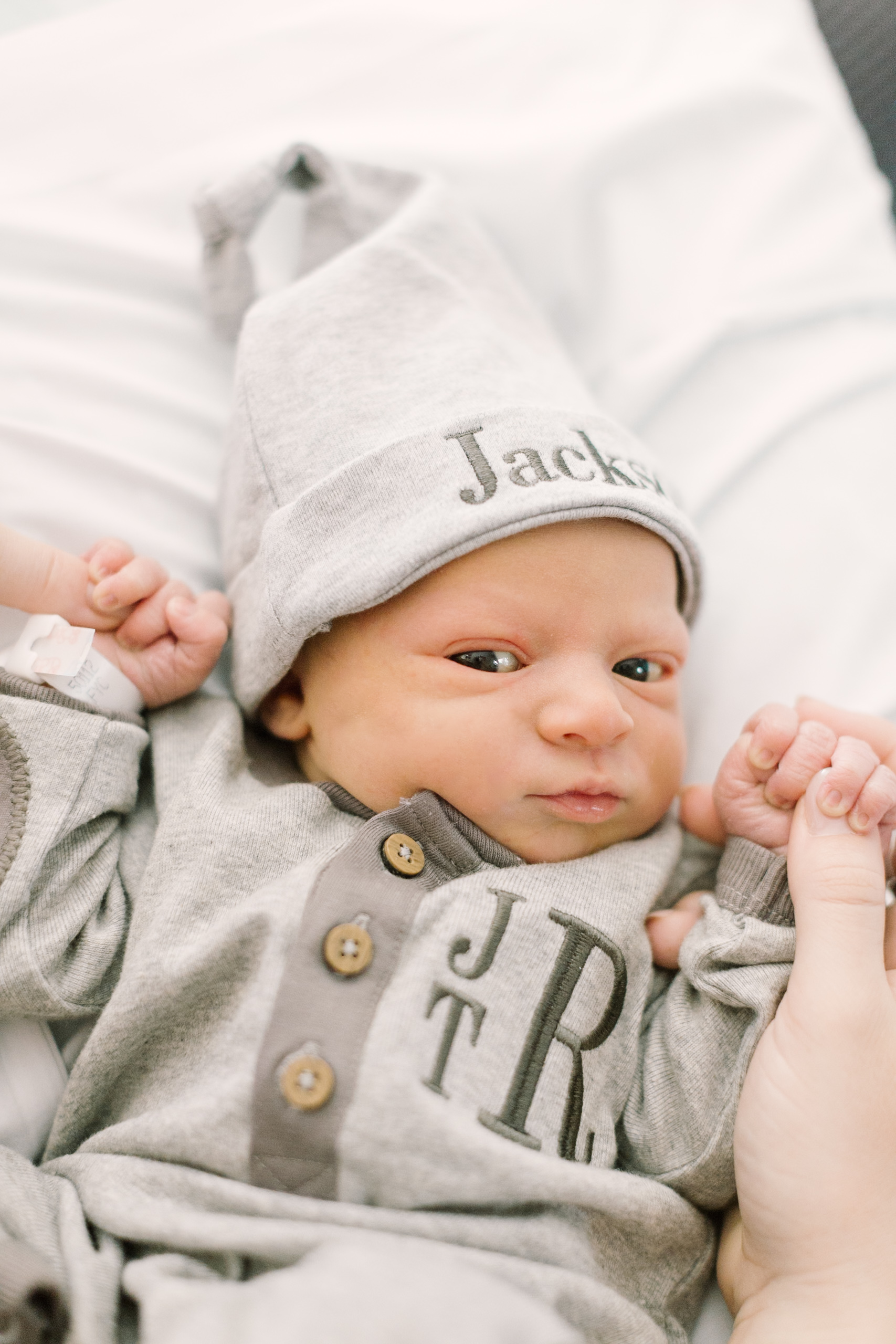 A look down on an awake newborn holding mom's thumbs on a bed in custom grey onesie and sleep cap before meeting babysitters in raleigh nc