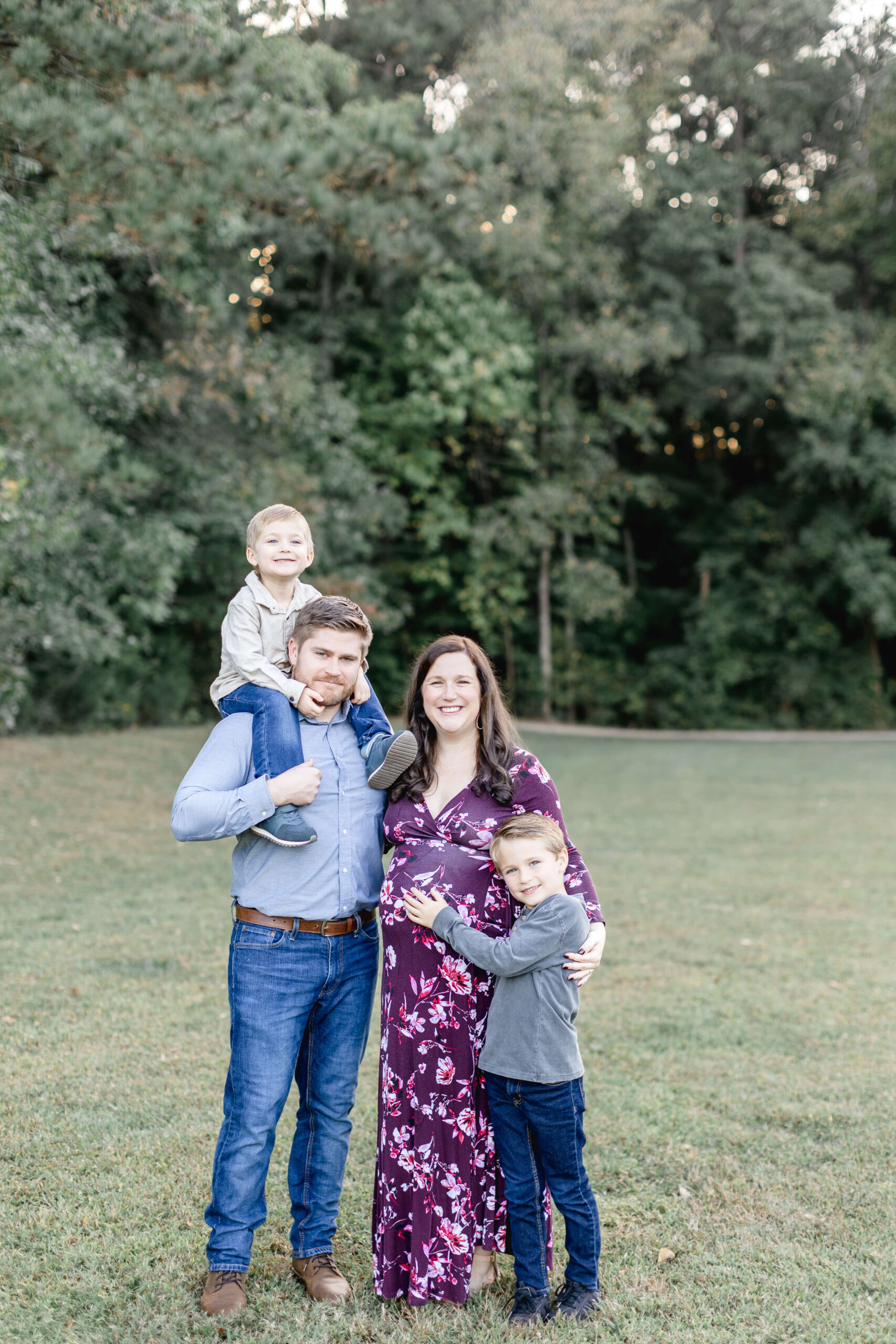 A smiling pregnant mom stands with her husband and two young sons in a park at sunset