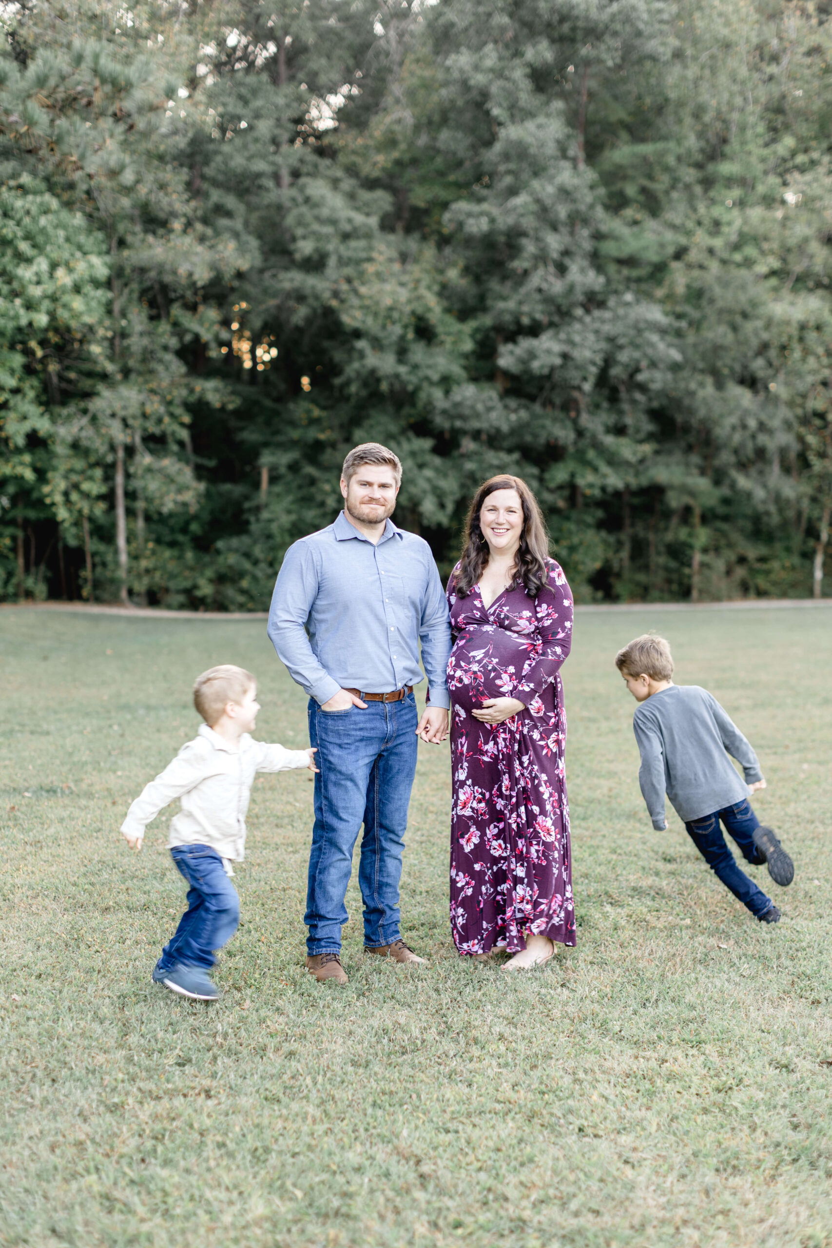 two toddler boys in jeans run circles around pregnant mom and dad standing in a park field after visiting a birth center in raleigh