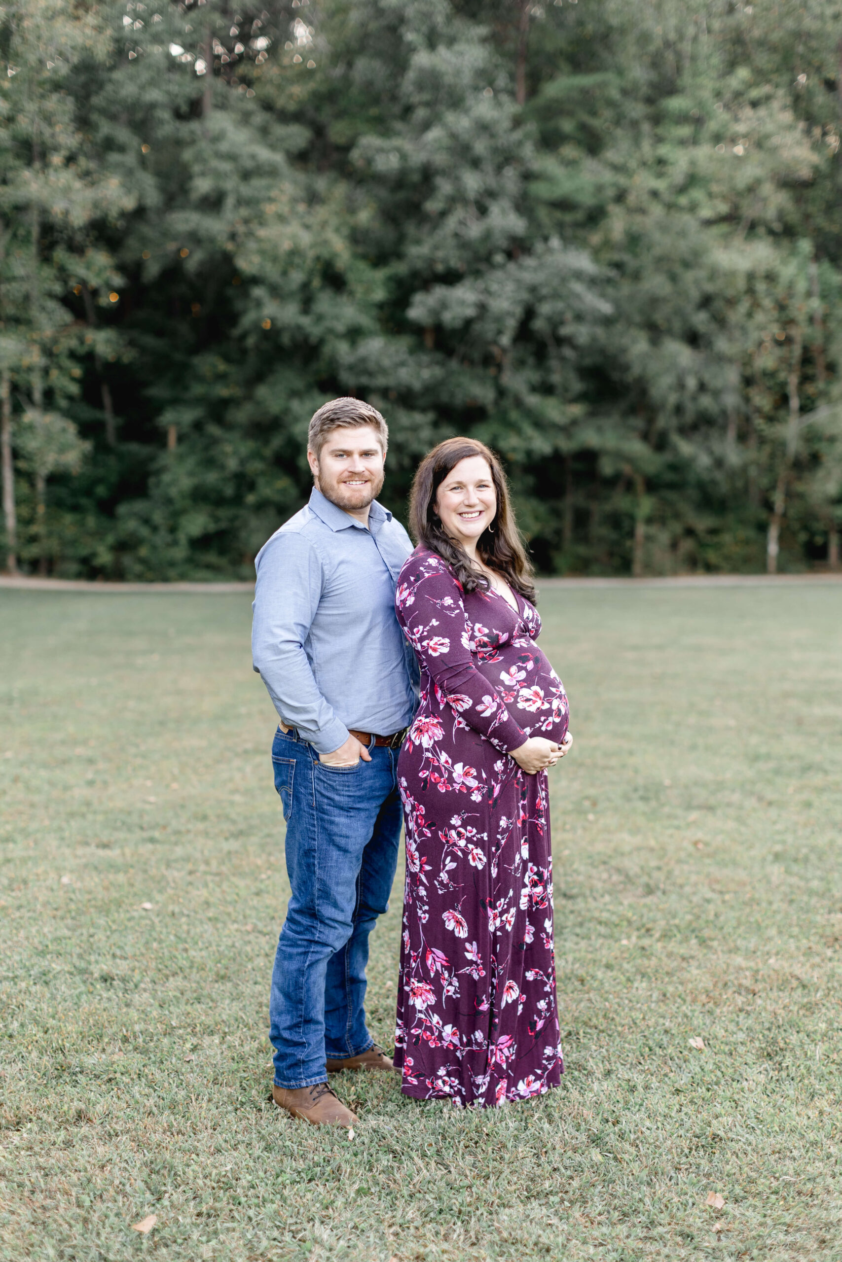 a happy expecting couple in blue and purple stand in a park field smiling after visiting a birth center in raleigh