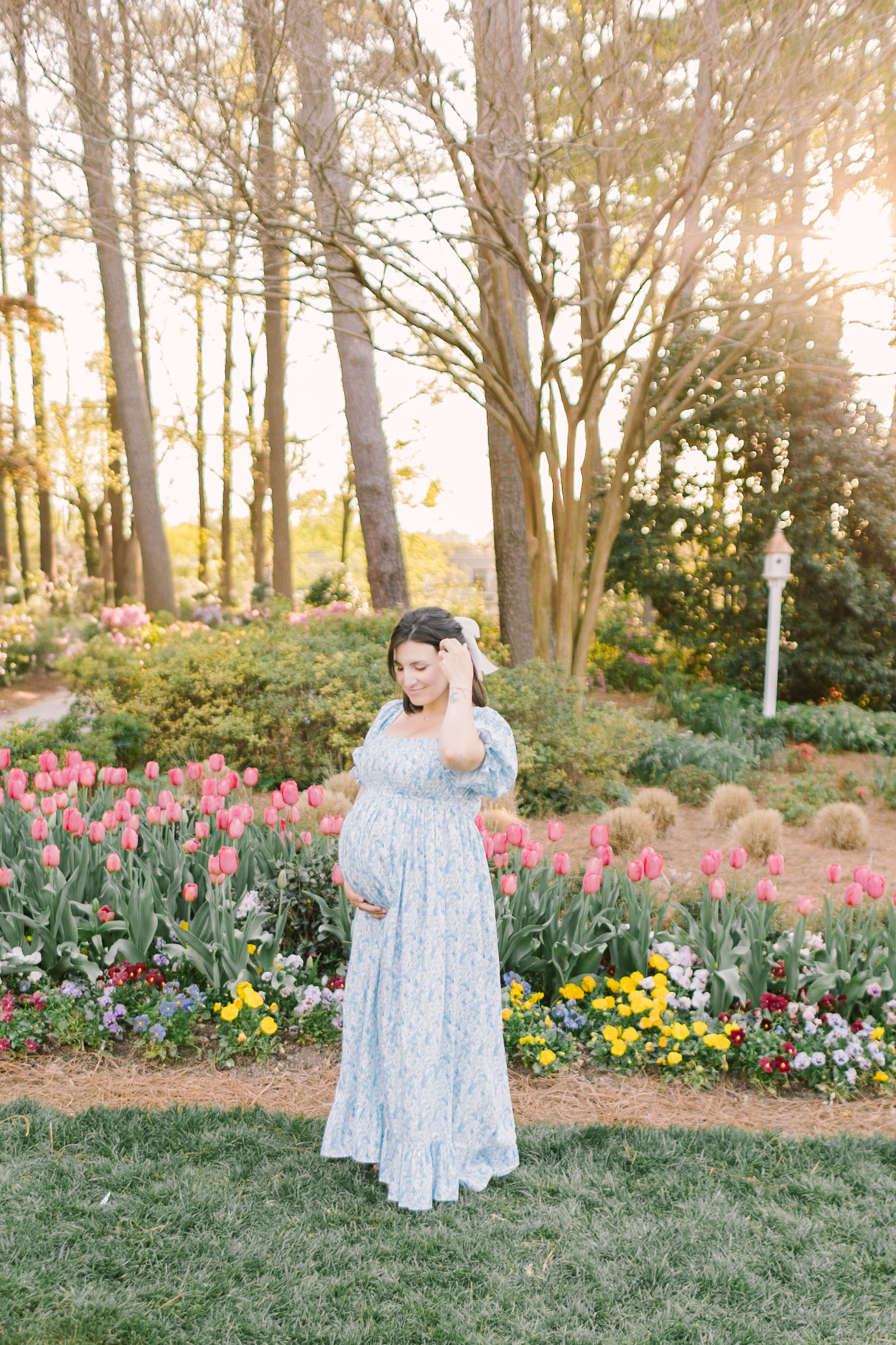 A happy mother to be pulls her hair behind her ear while standing in a colorful garden at sunset in a blue maternity gown