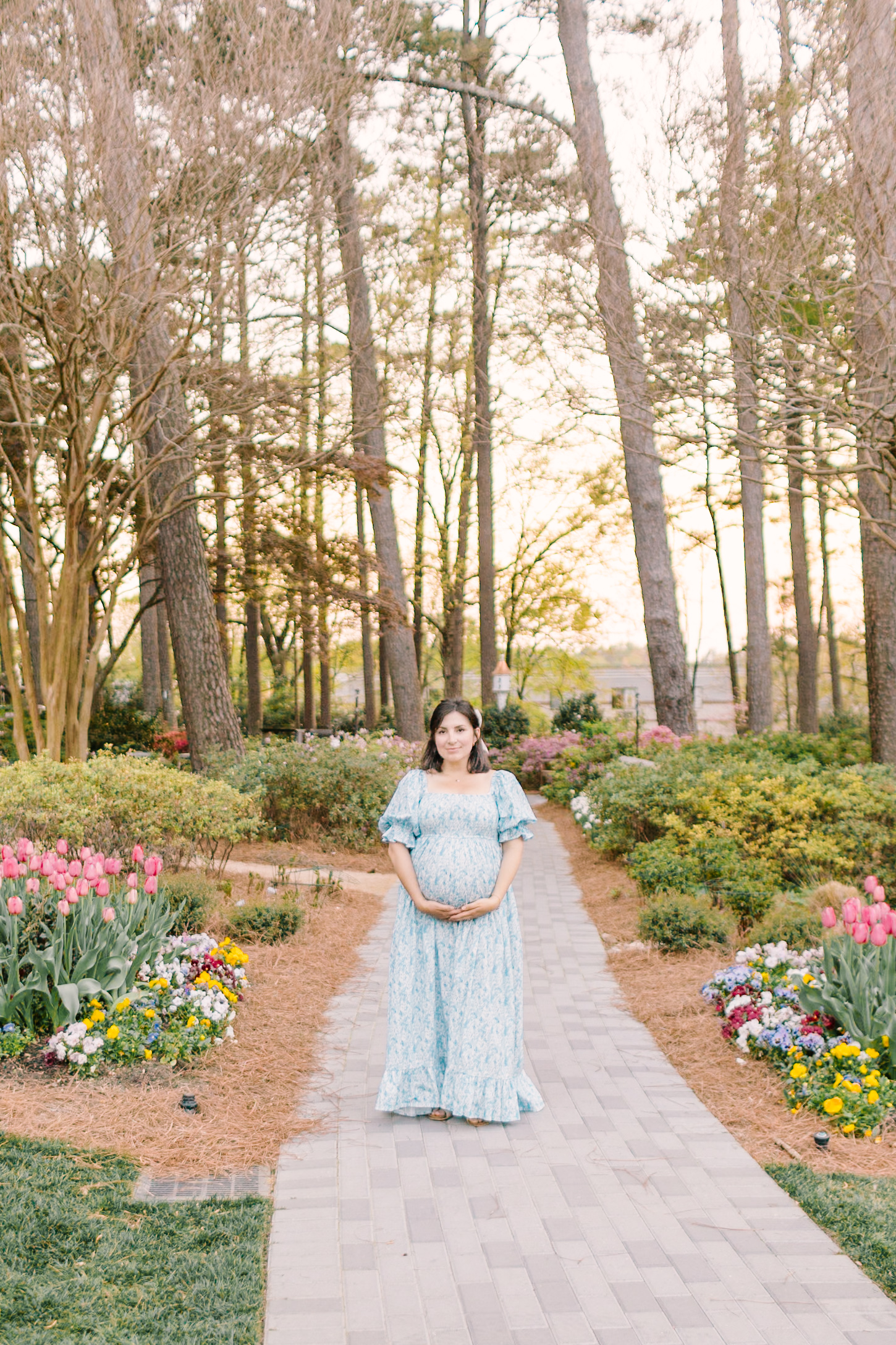 A mom to be in a blue maternity gown walks in a vibrant garden at sunset with hands under her bump thanks to ivf in raleigh