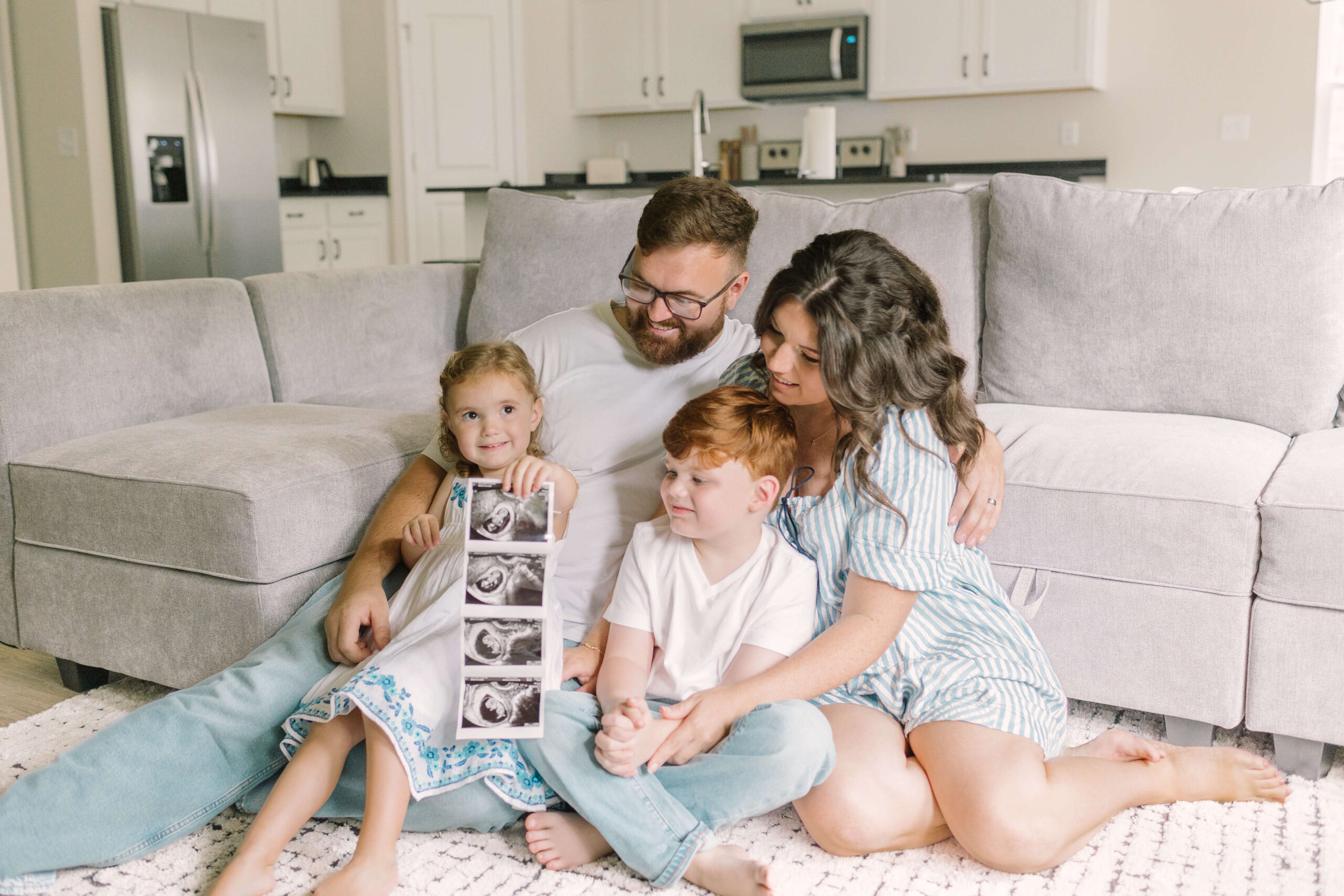 A toddler girl holds a sonogram while sitting in dad's lap on the floor against a couch with older brother