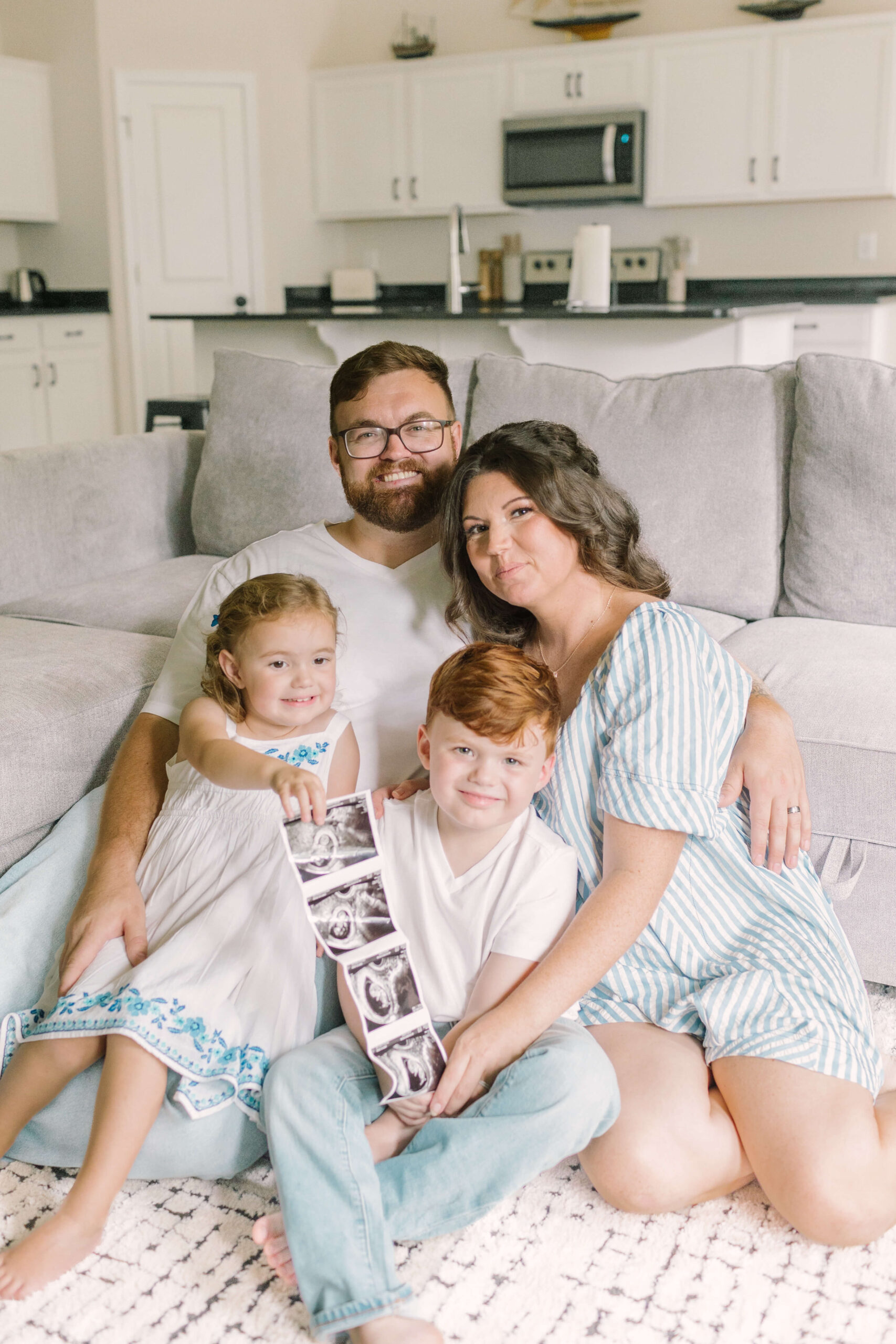a mom and dad sit on the floor with their toddler son and daughter in their laps holding a sonogram