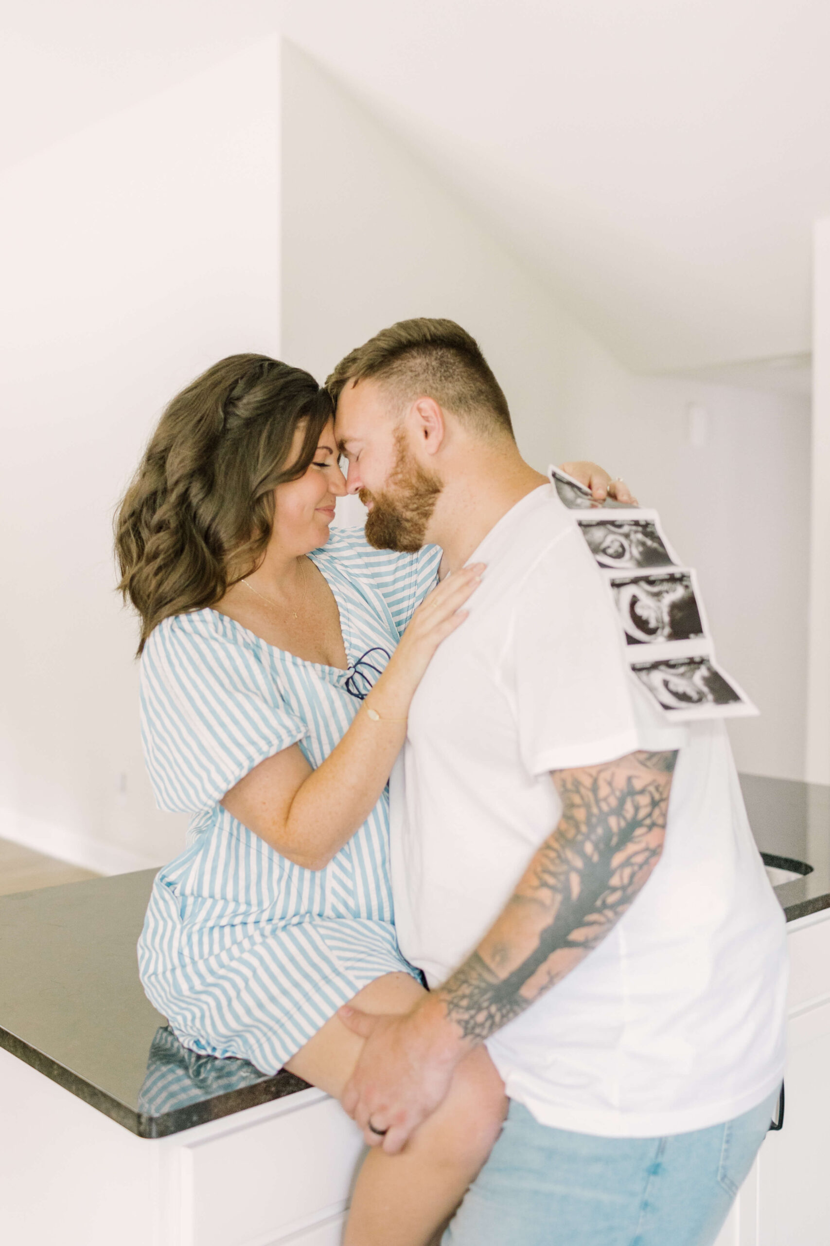A happy pregnant couple touch foreheads on the kitchen counter holding a sonogram after meeting midwives in raleigh