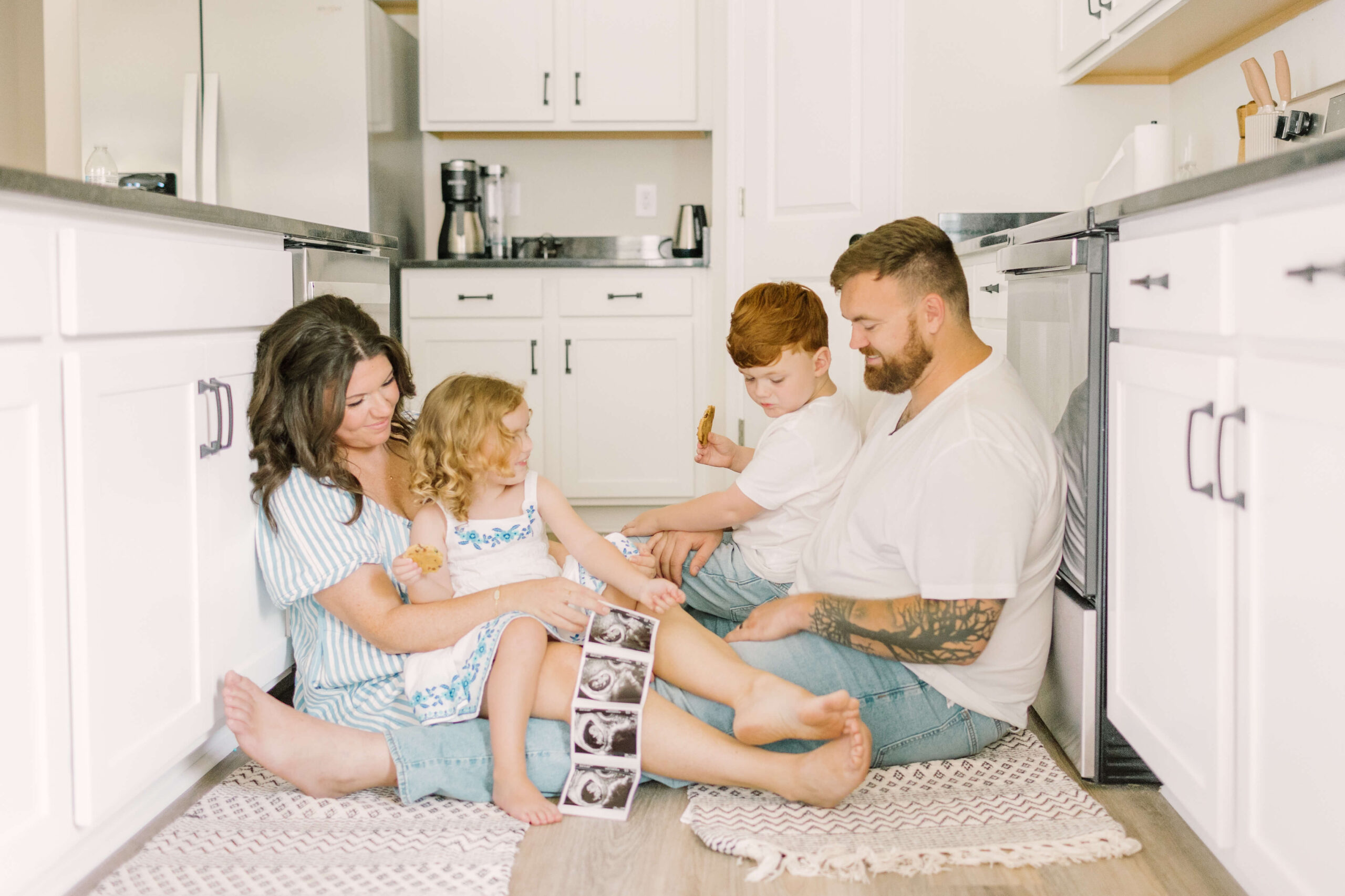 A mom and dad sit on the kitchen floor with their two toddlers in their laps holding a sonogram after finding midwives in raleigh