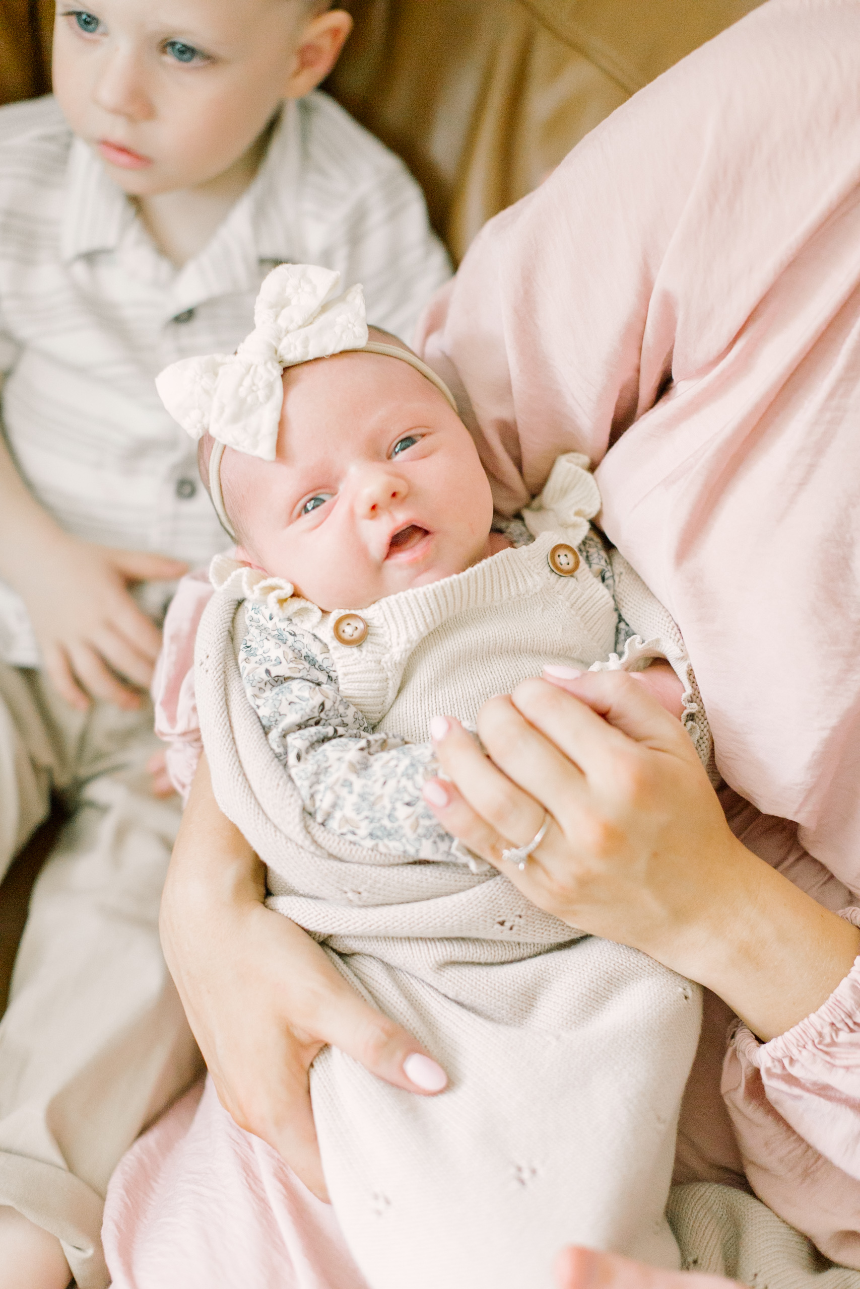 an awake newborn baby lays in mom's lap in a blanket and dress with overalls on a leather couch