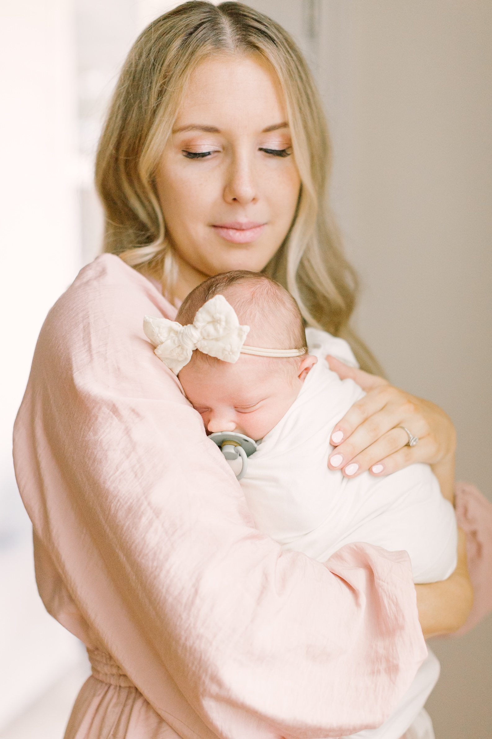 a new mother in a pink dress stands cradling her sleeping newborn against her chest in a white swaddle and bow