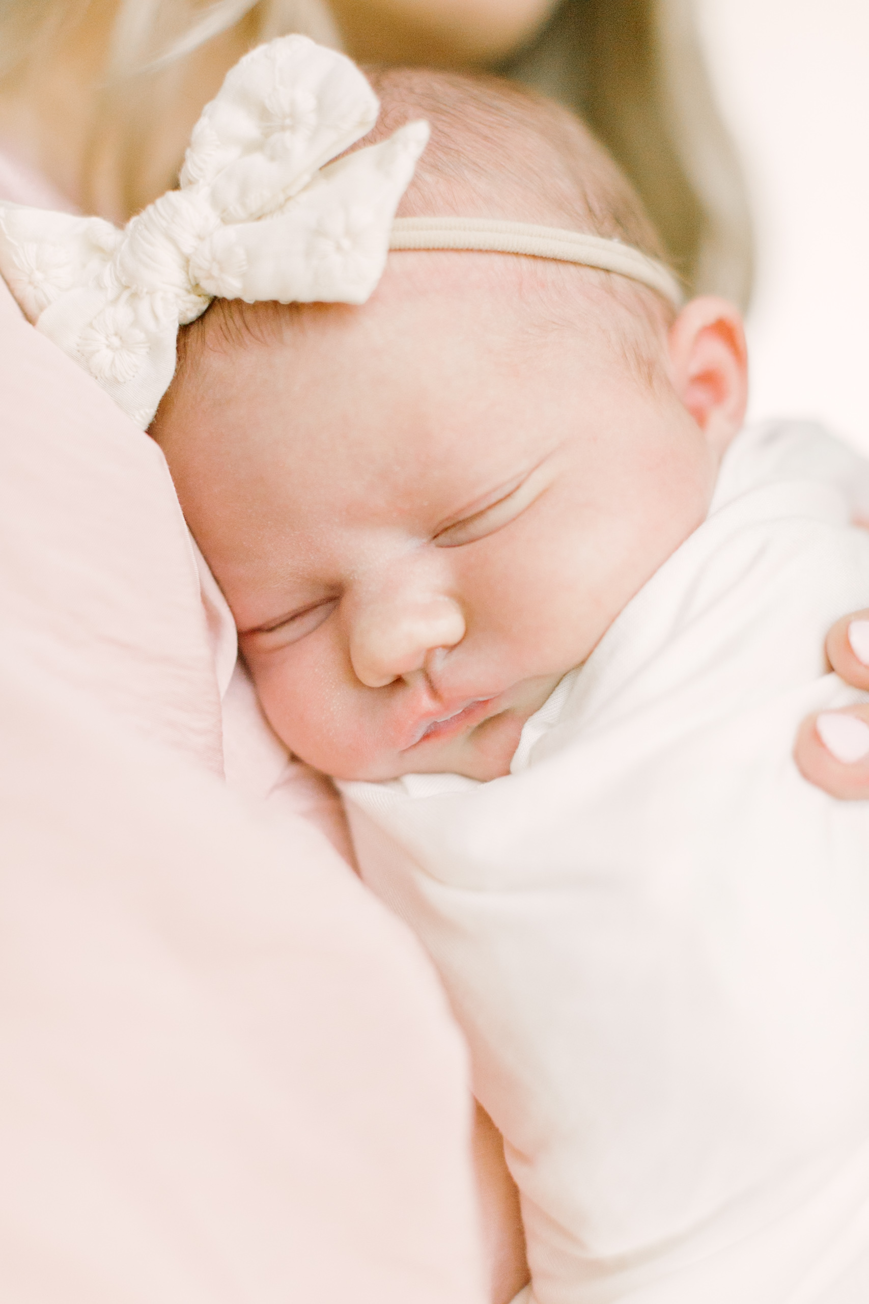 A newborn baby in a white swaddle and matching bow sleeps against mom's shoulder in a pink dress thanks to an obgyn in raleigh