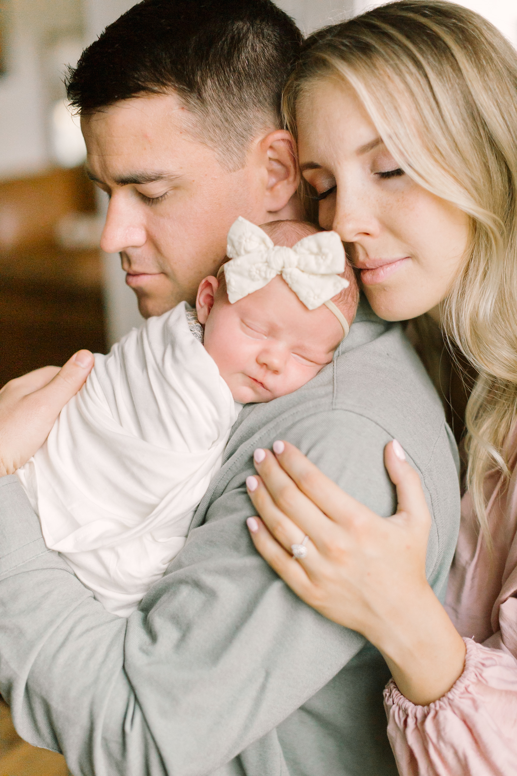 Happy new parents snuggle while standing with their newborn girl sleeping on dad's shoulder thanks to an obgyn in raleigh