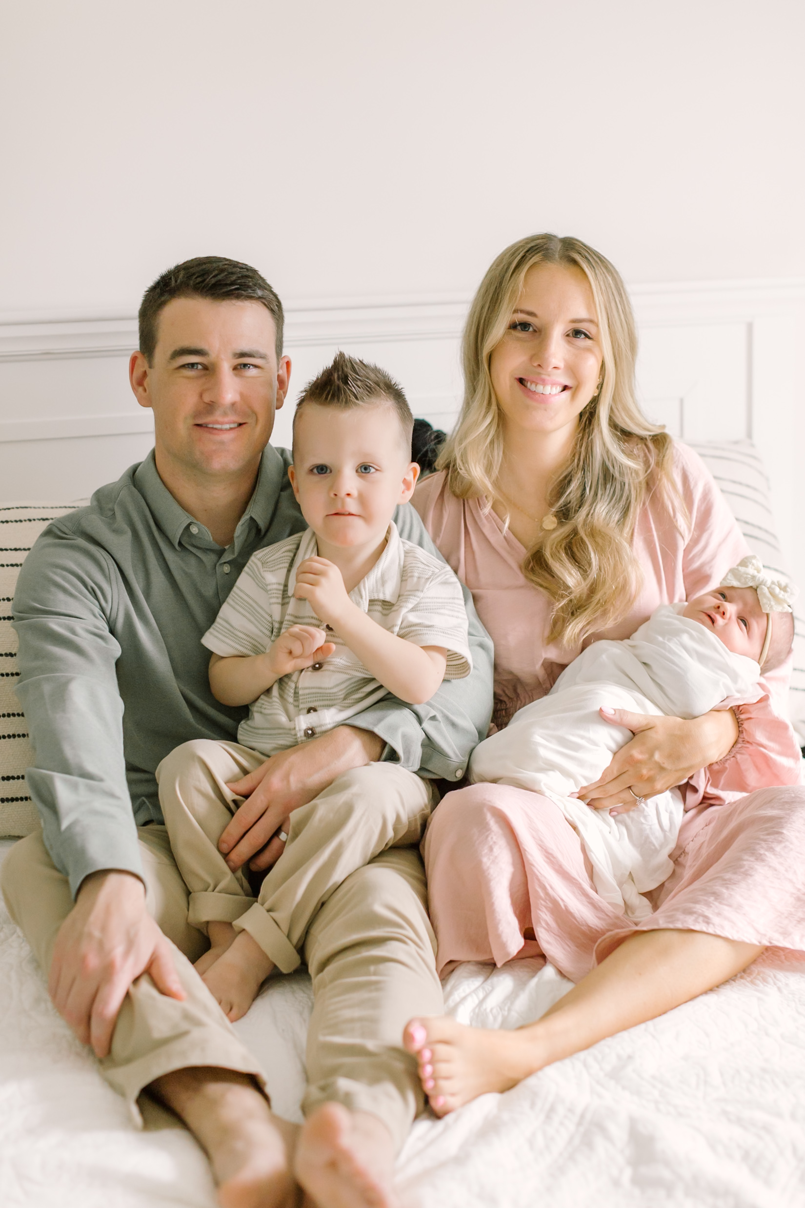 smiling mom and dad sit on a bed in a pink dress and blue shirt with their toddler son and newborn in their laps thanks to an obgyn in raleigh