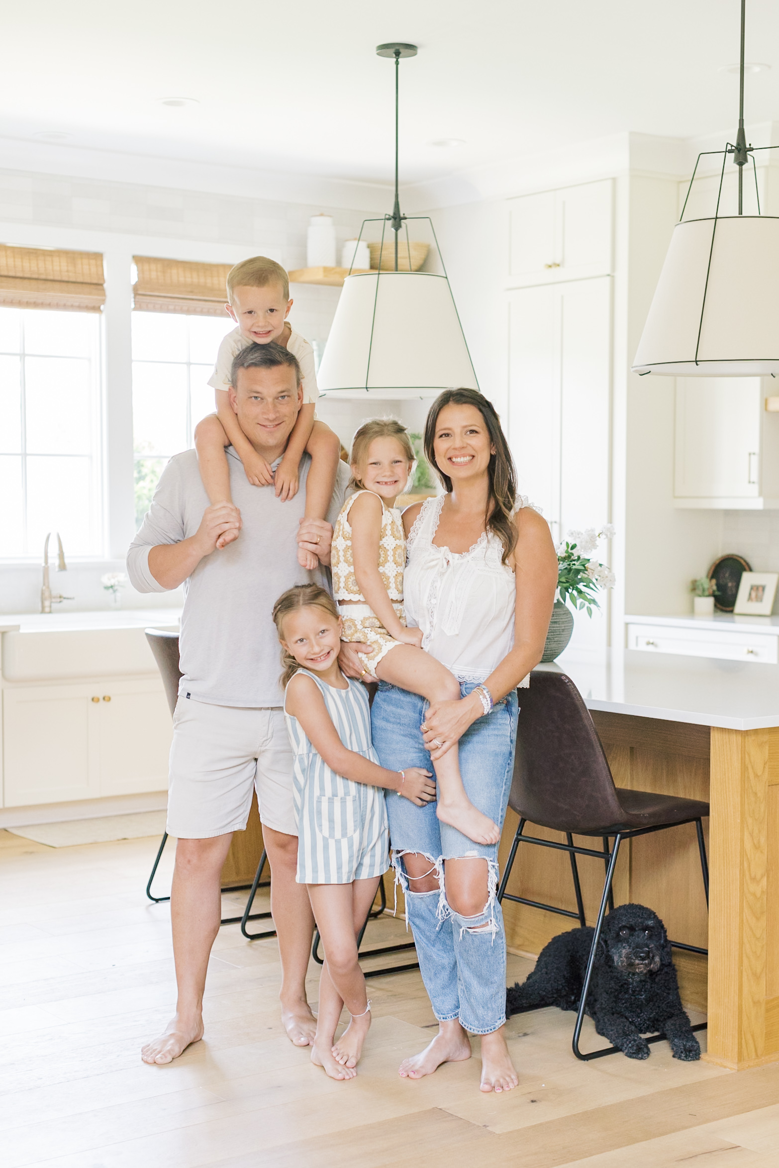 A mom and dad stand in their kitchen with their three children on their shoulders, hips and hugging their leg
