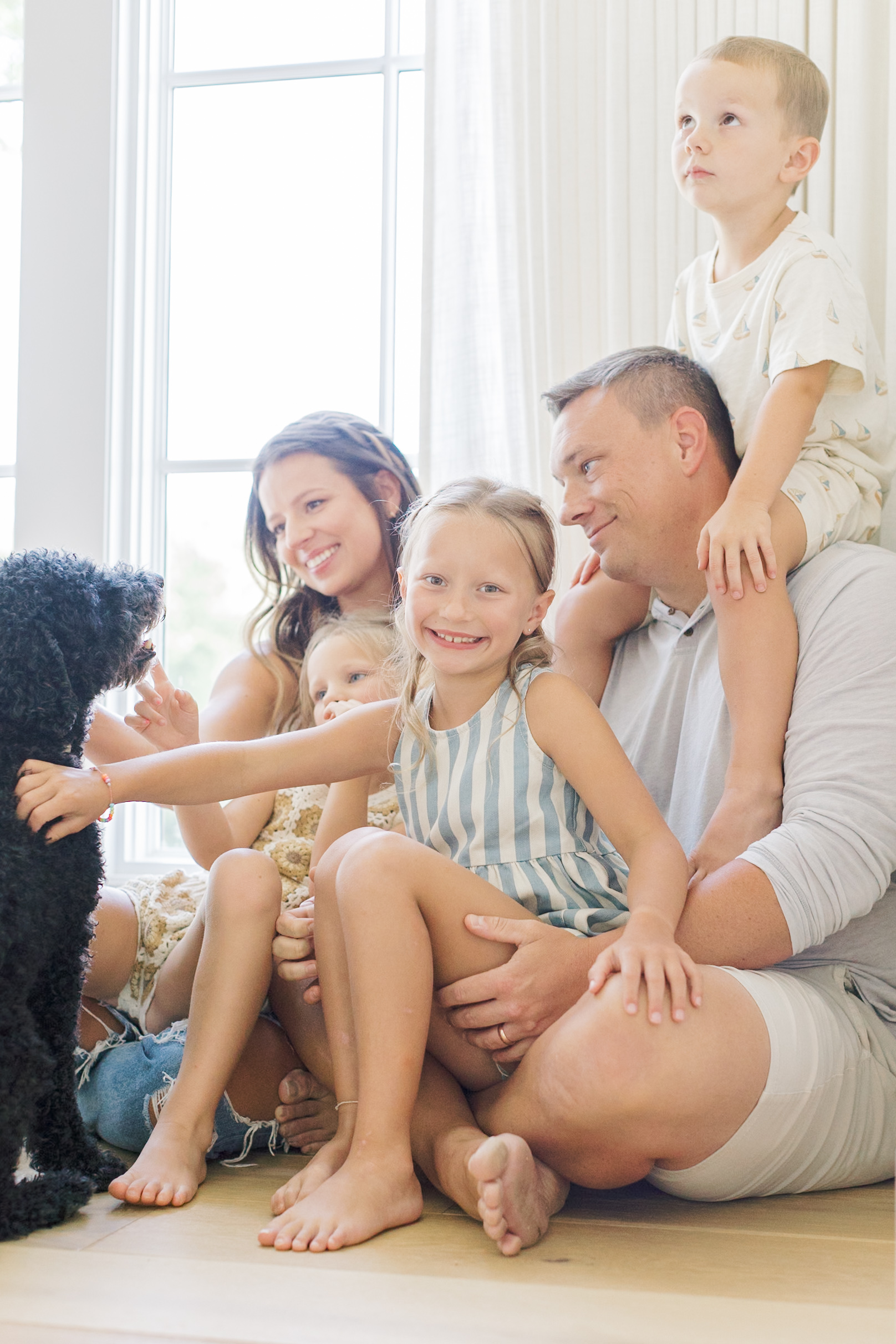 a happy toddler girl sits in dad's lap on the floor as their family of five play with a black dog under a window after some parenting classes in raleigh nc