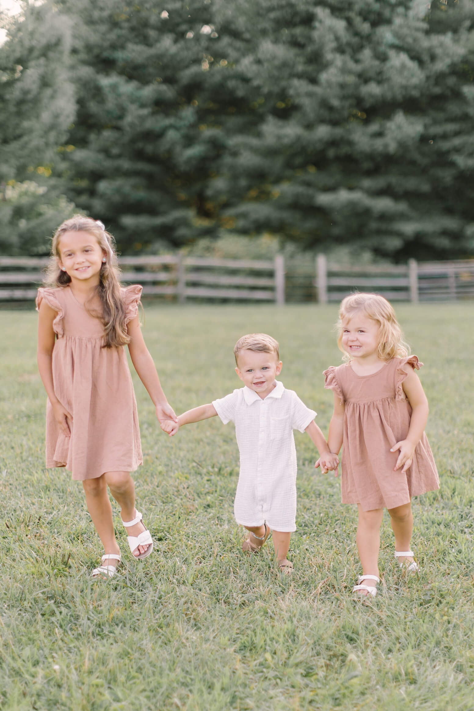 a toddler boy in a white onesie walks holding hands with his two toddler sisters in a park at sunset with big smiles