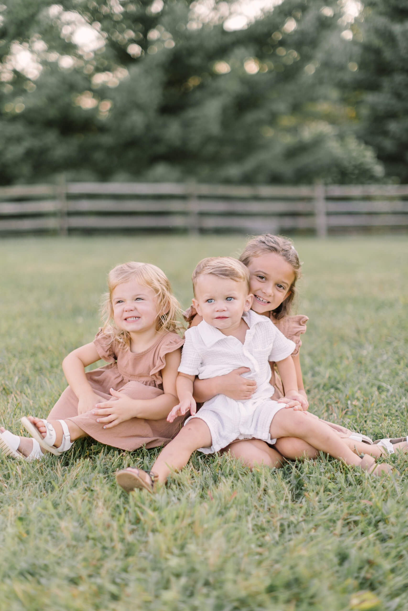 a toddler boy in a white onesie sits in his sisters lap with matching sister next to them in a park lawn at sunset