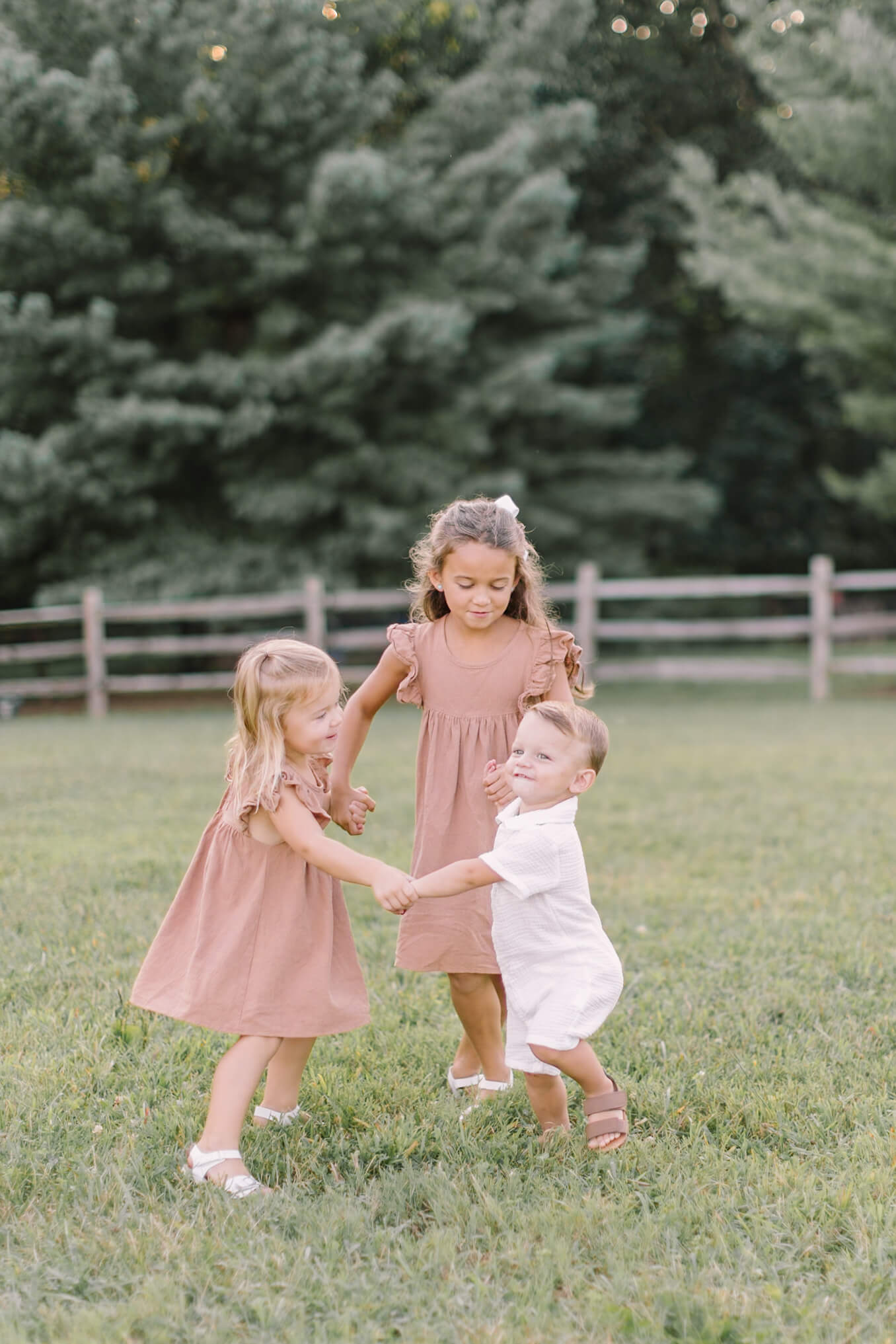 Two toddler sisters in matching brown dresses dance in a circle holding hands with younger brother in a field in a white onesie all with big smiles after visiting preschools in raleigh