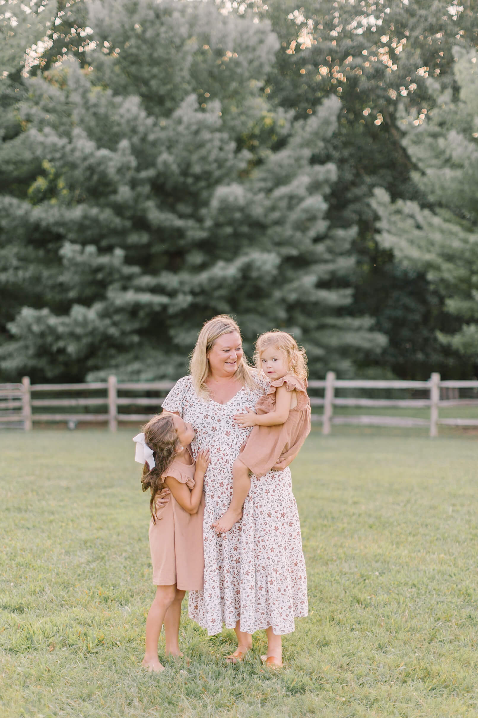 a mother in a white floral print dress smiles while holding a toddler girl on her hip in a brown dress matching big sister's hugging mom in a park at sunset after visiting preschools in raleigh