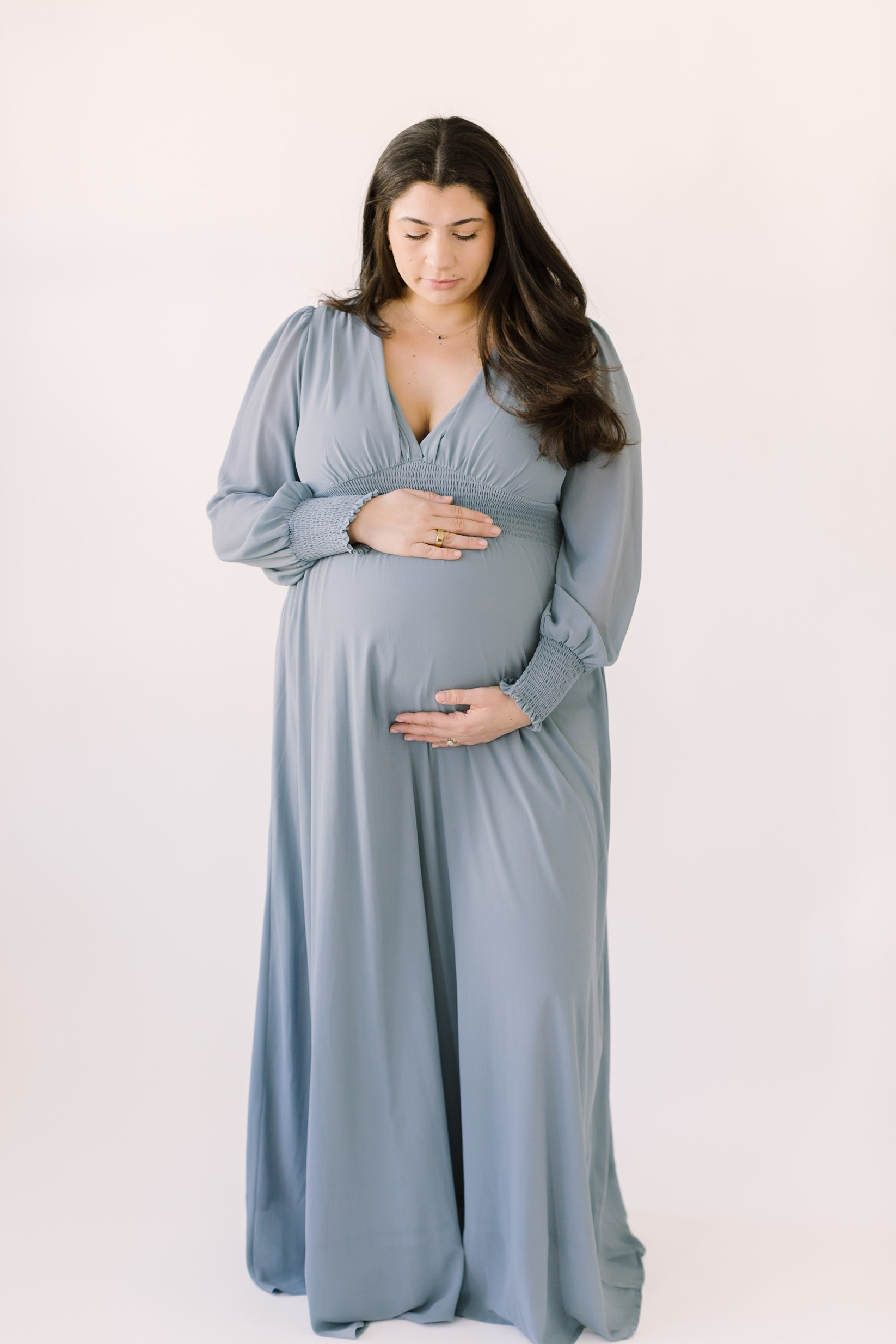 A mom to be looks down to her bump in a blue maternity gown with hands on top and bottom