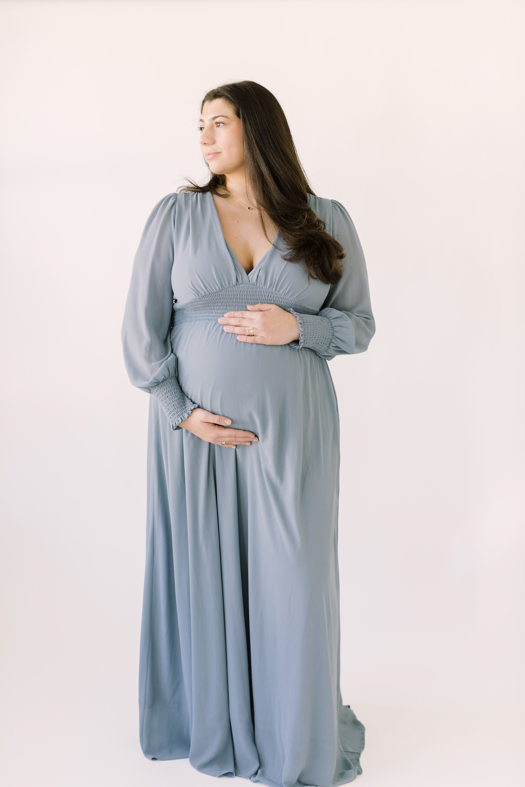 A happy expecting woman in a blue maternity dress stands in a studio looking over her shoulder after visiting baby shower venues in raleigh nc