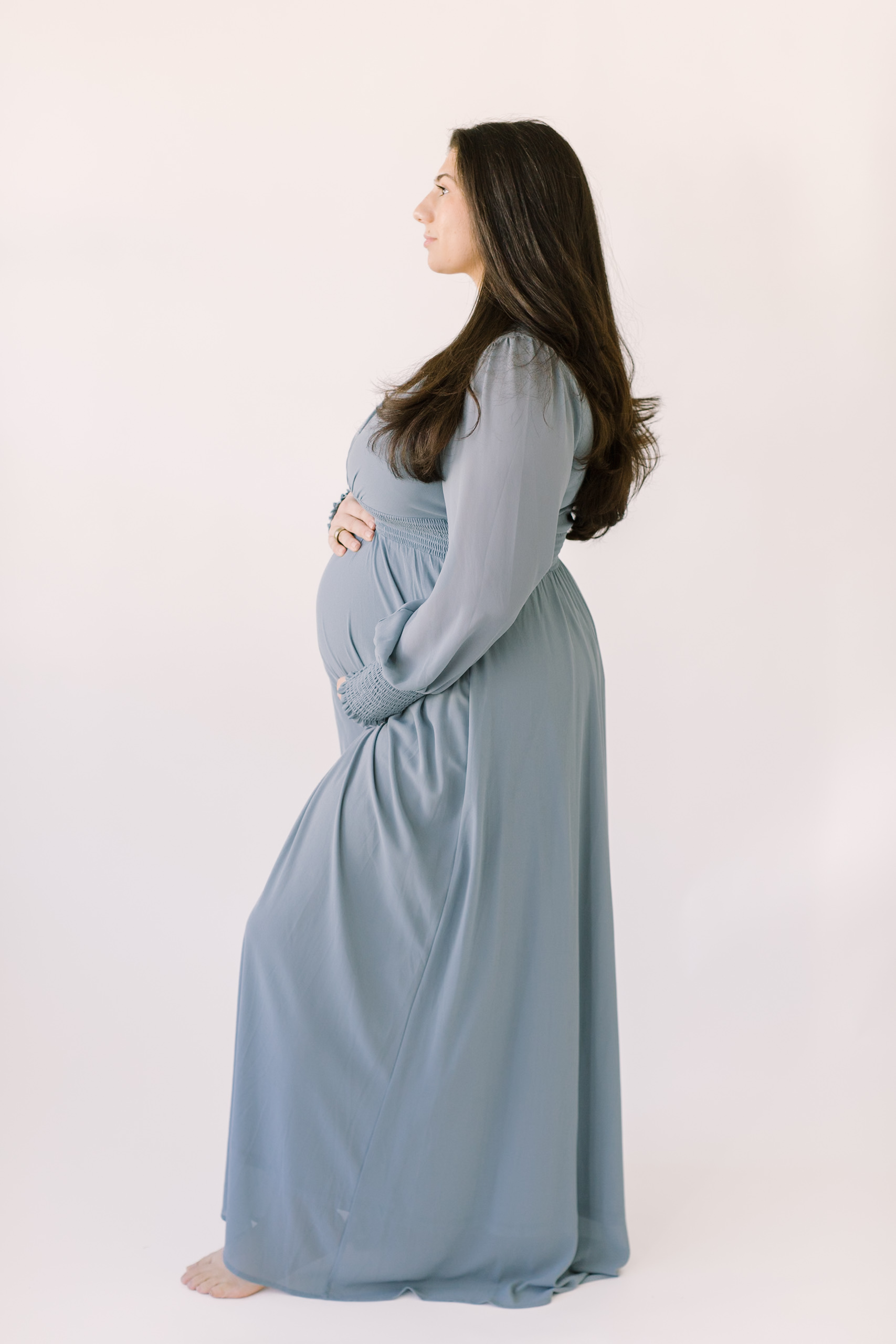 A mother to be gazes out a window in a studio in profile while holding her bump in a blue maternity gown after visiting baby shower venues in raleigh nc