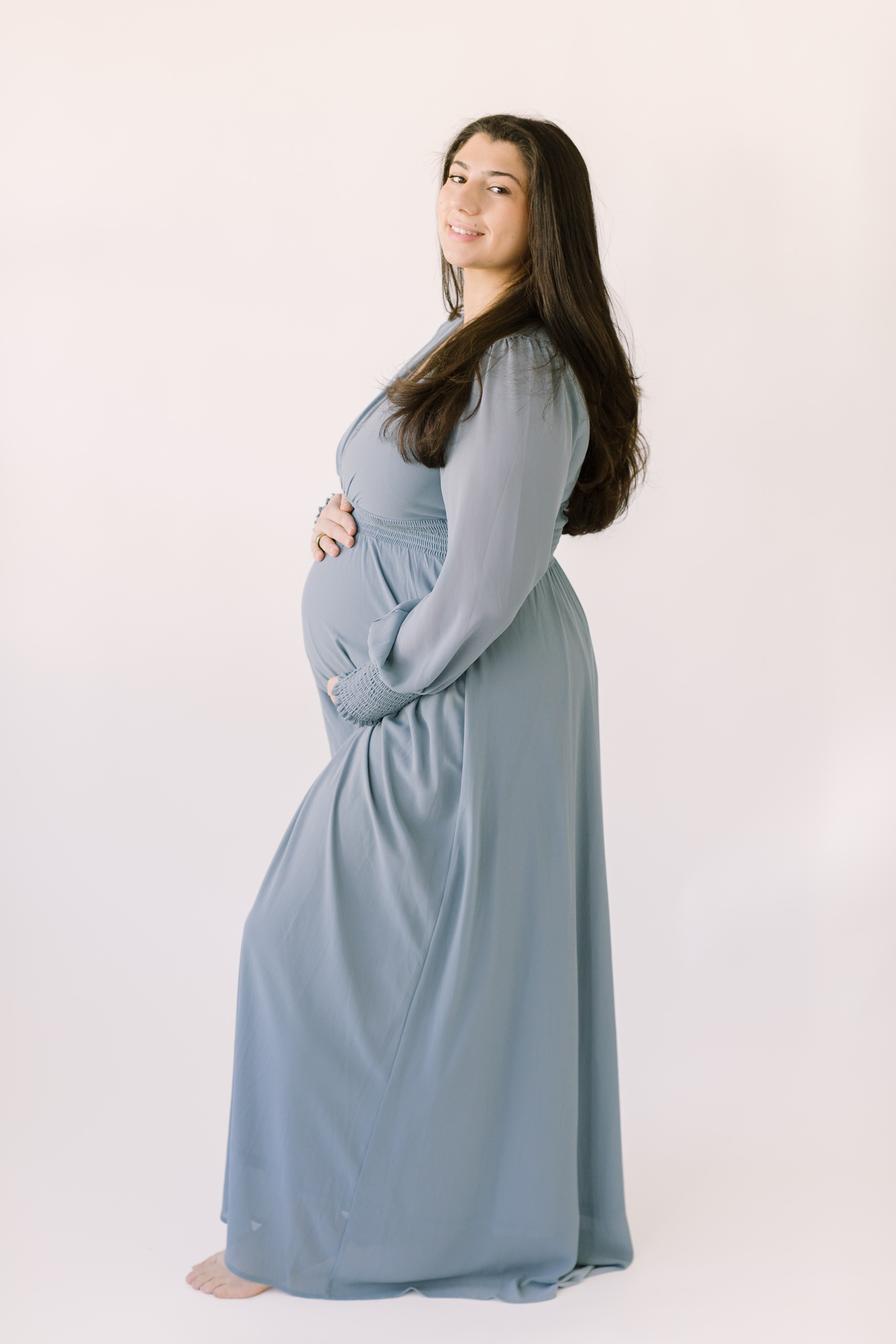 A pregnant woman stands in a studio in a blue maternity gown smiling over her shoulder with hands on top and bottom of bump after visiting baby shower venues in raleigh nc