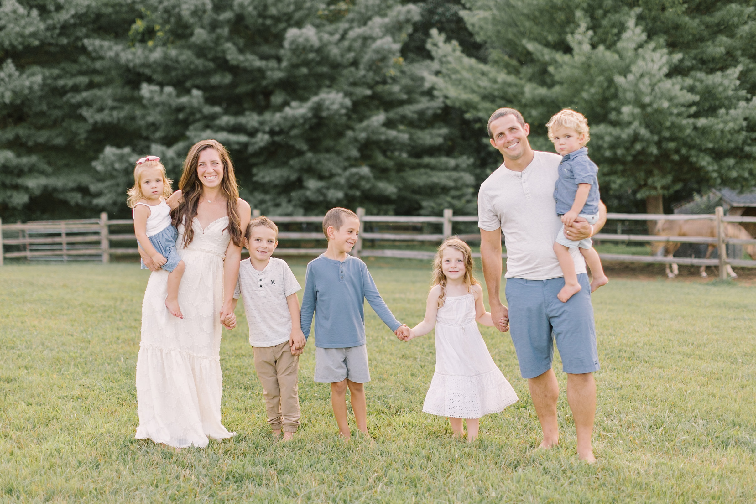 A smiling mom and dad walk holding hands and carrying their five young children in a pasture at sunset in blue and white outfits