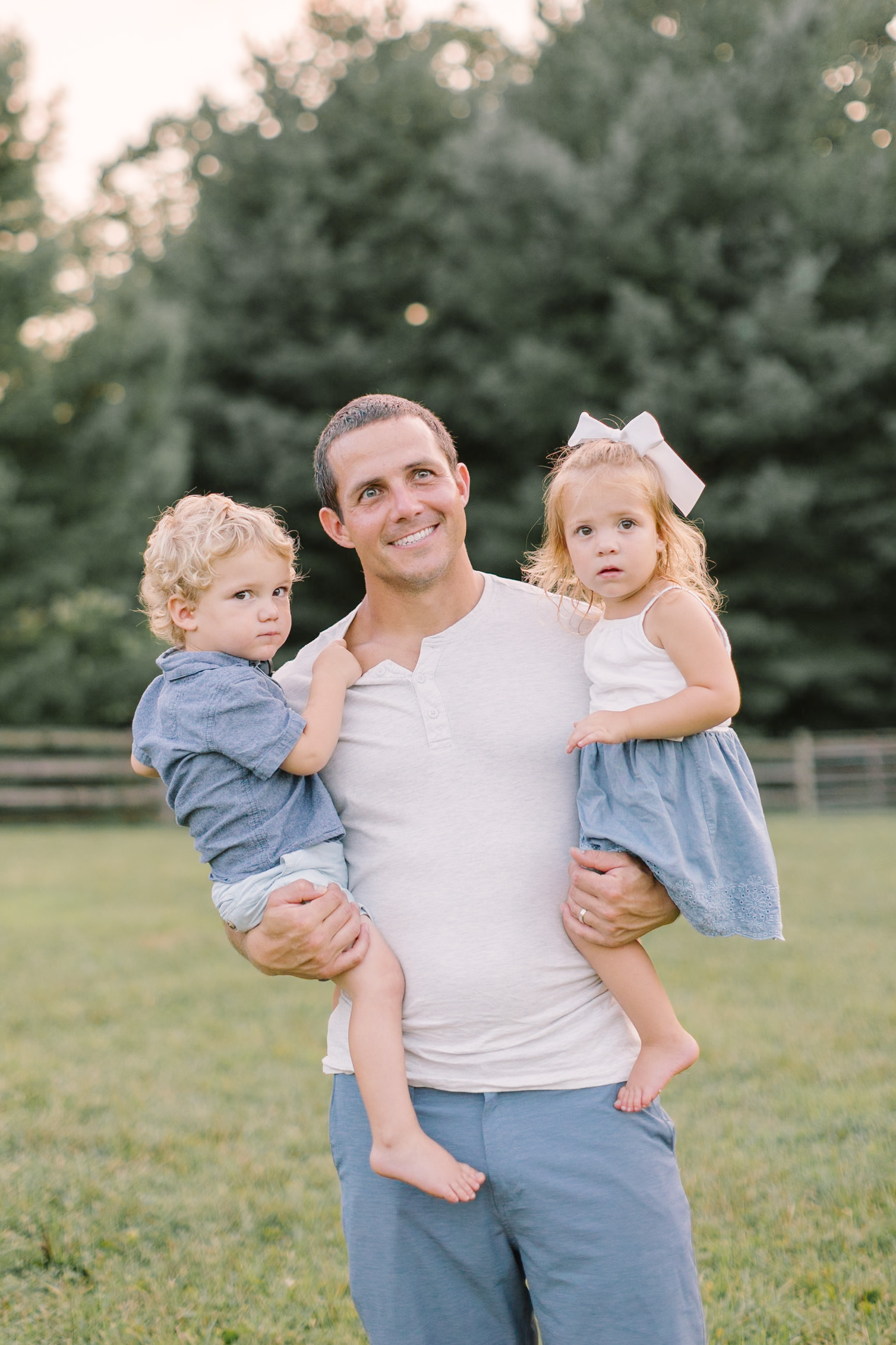 A dad in a white shirt holds his twin toddlers in his arms while standing in a pasture at sunset after visiting children's boutiques in Raleigh, NC