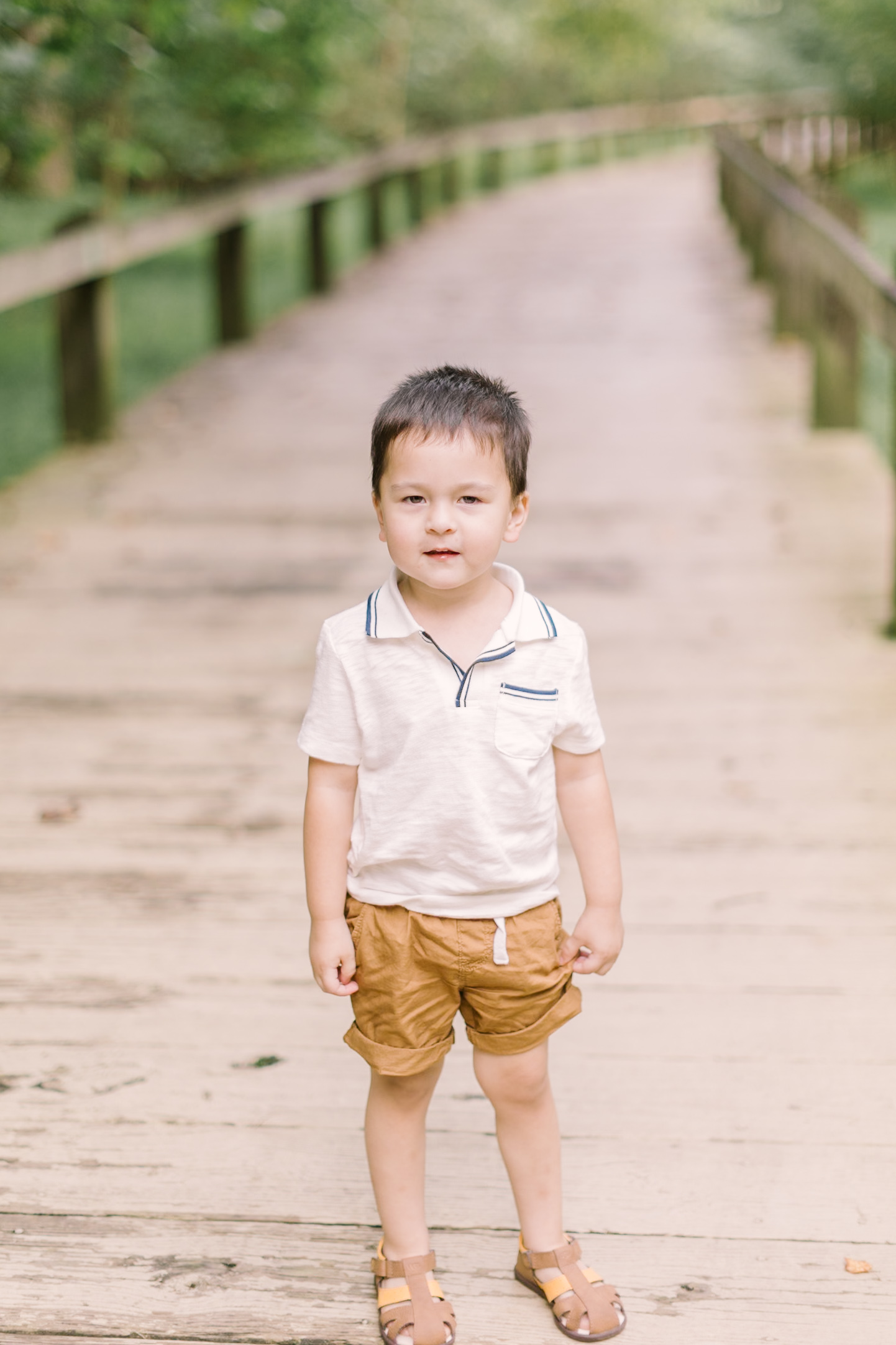 A toddler boy in khaki shorts and white shirt stands in the middle of a boardwalk