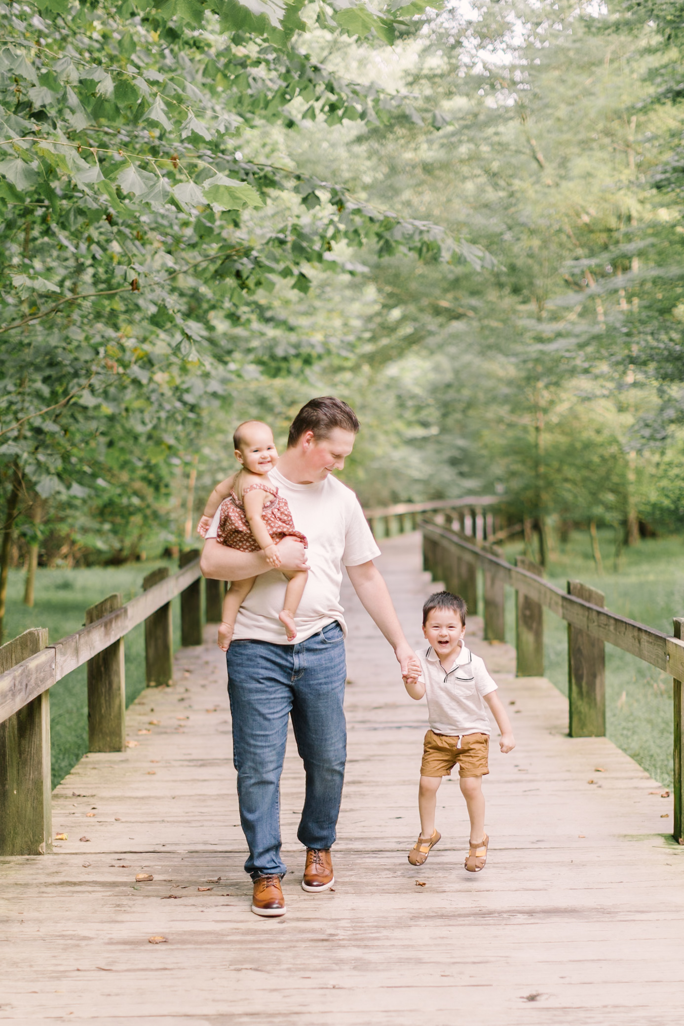 A happy dad in a white shirt and jeans walks his toddler son by the hand down a boardwalk with his infant in one arm