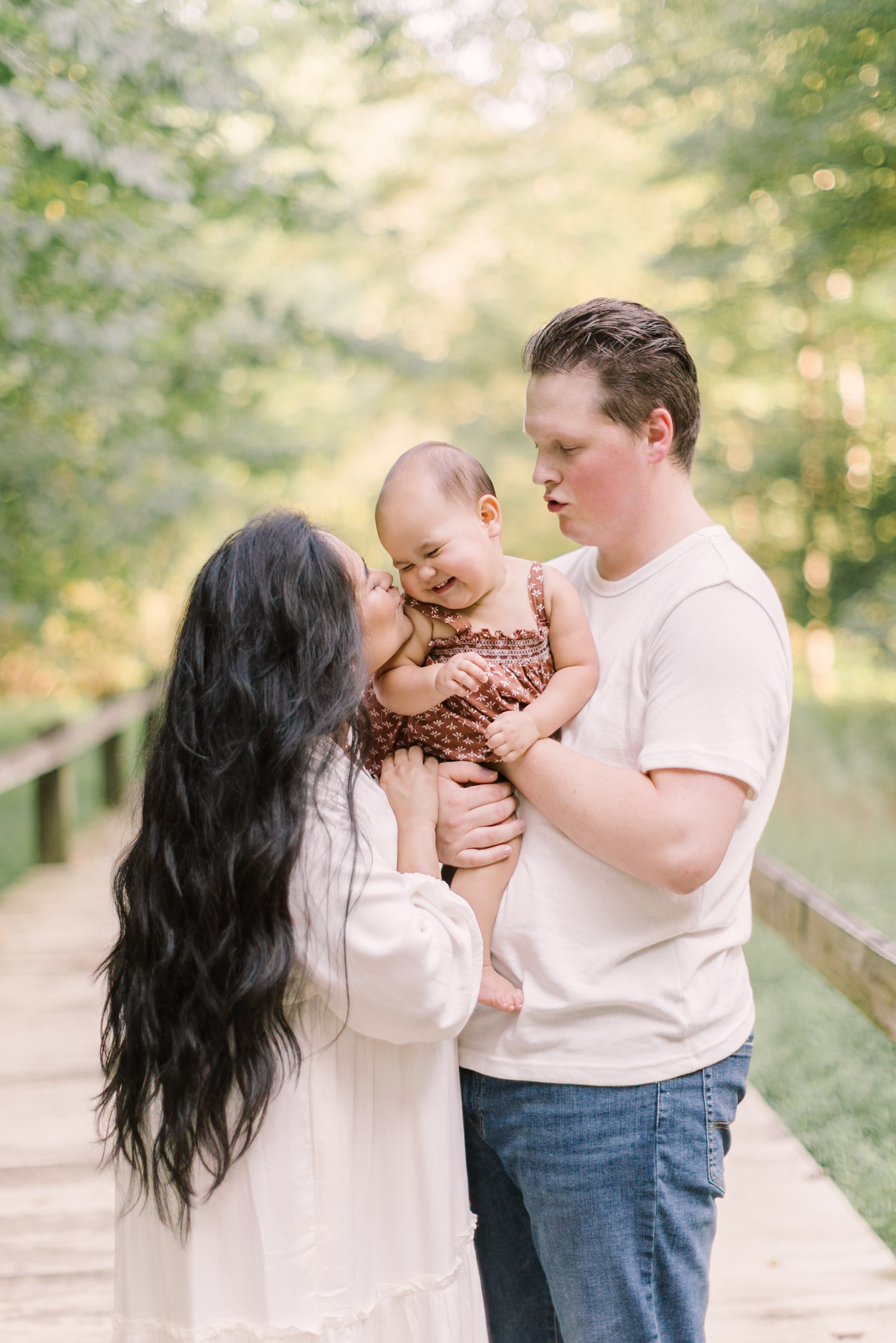 A mom in a white dress leans in to kiss her giggling infant in dad's arms while standing on a boardwalk after visiting toy stores in raleigh