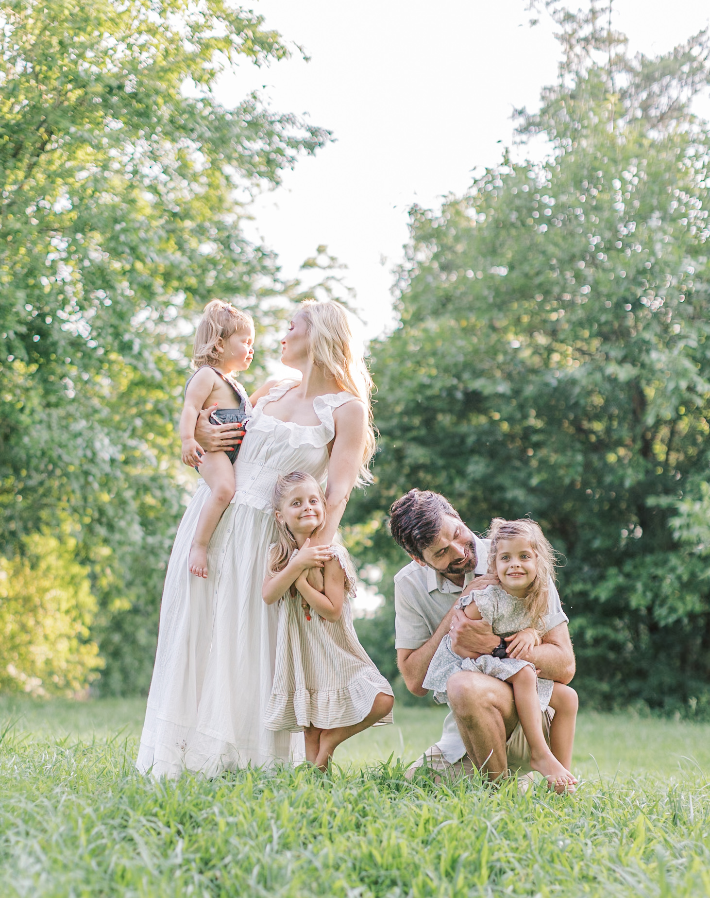 Three toddler sisters are hugged and held by their mom and dad in a park lawn