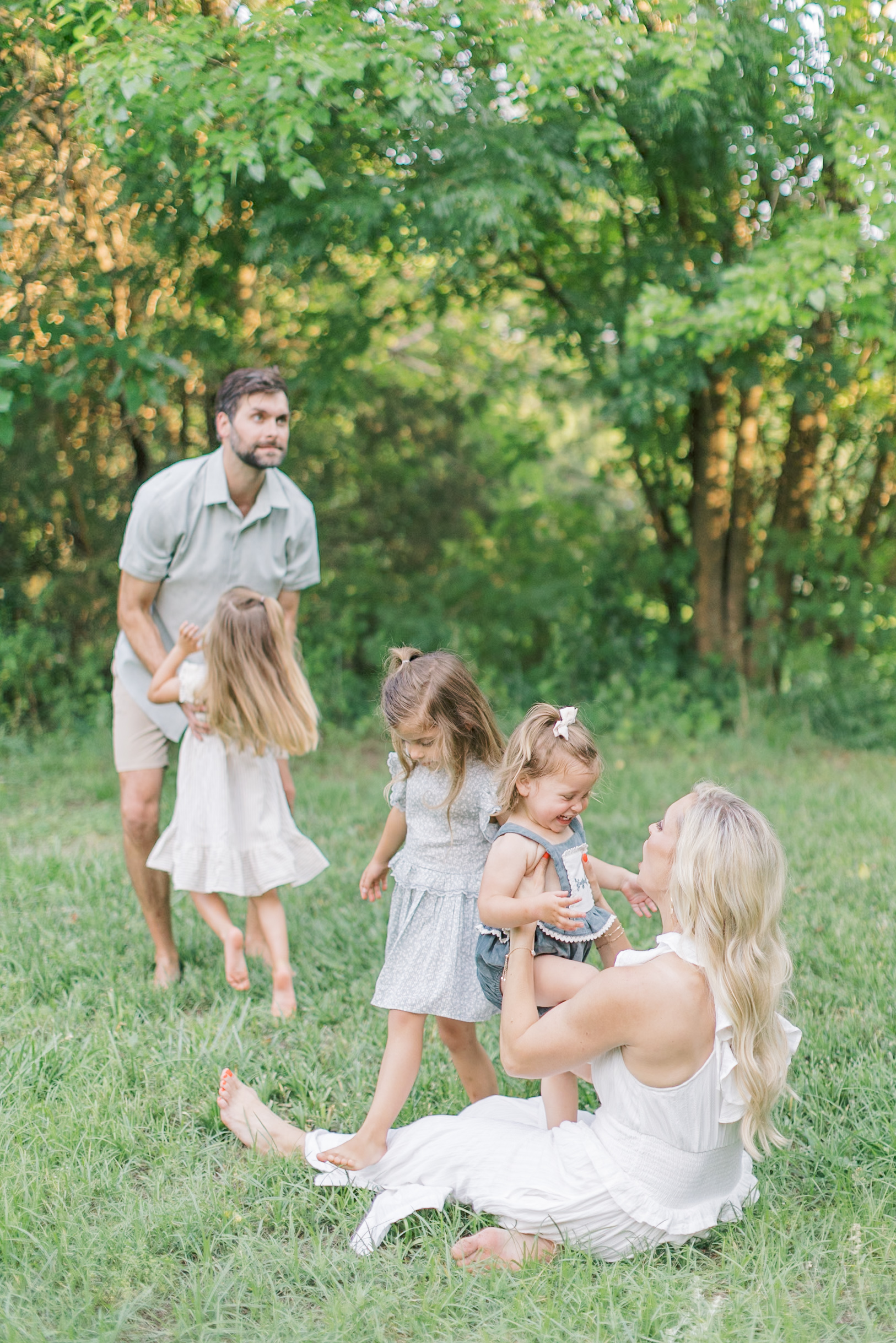 A mom and dad play with their three toddler daughters in a park lawn barefoot in white and blue at sunset after finding great dance studios in Raleigh, NC