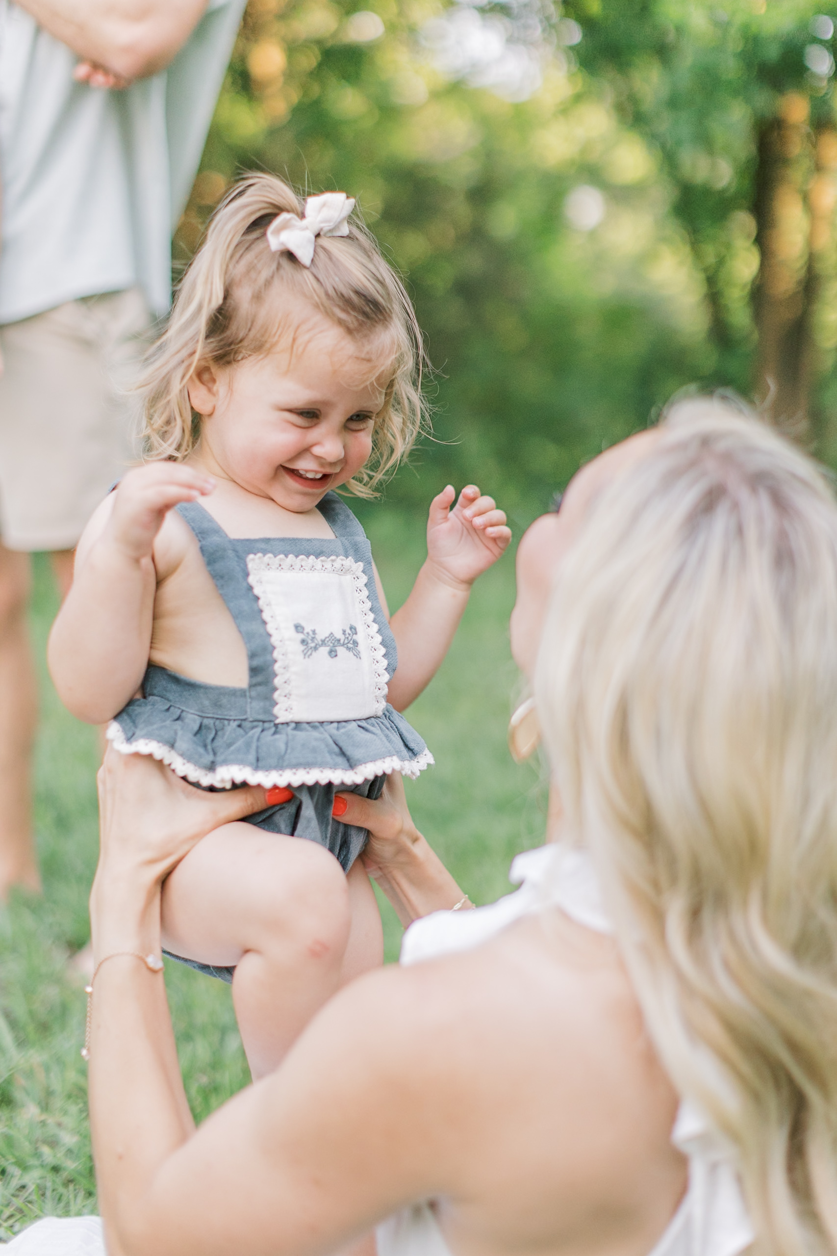 A toddler girl giggles while playing with mom in a park lawn in a blue onesie overalls