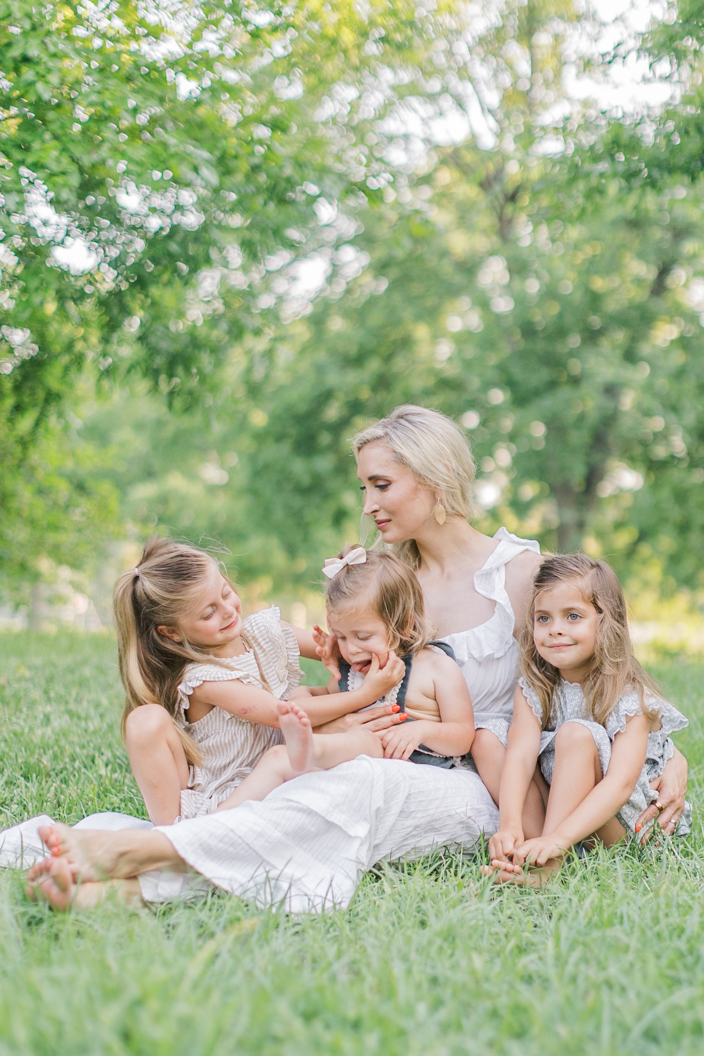 A mom in a white dress sits in a park lawn playing with three toddler girls in her lap after visiting dance studios in Raleigh, NC