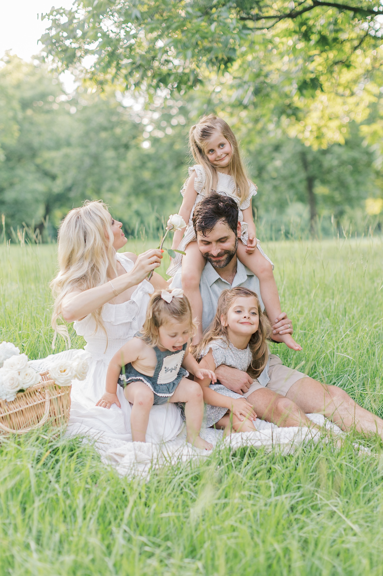 A mom and dad play with their three toddler daughters in a park lawn during a picnic after visiting dance studios in Raleigh, NC