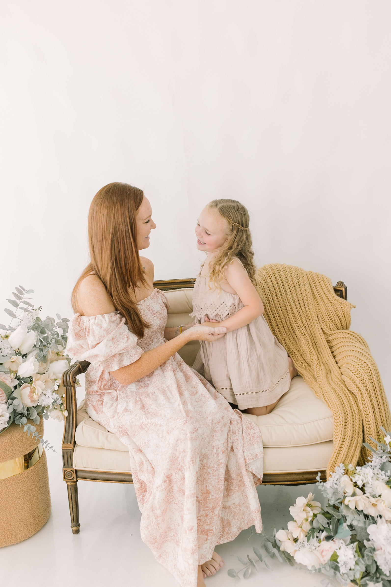 A toddler girl kneels on a bench with mom while holding hands and chatting in a studio