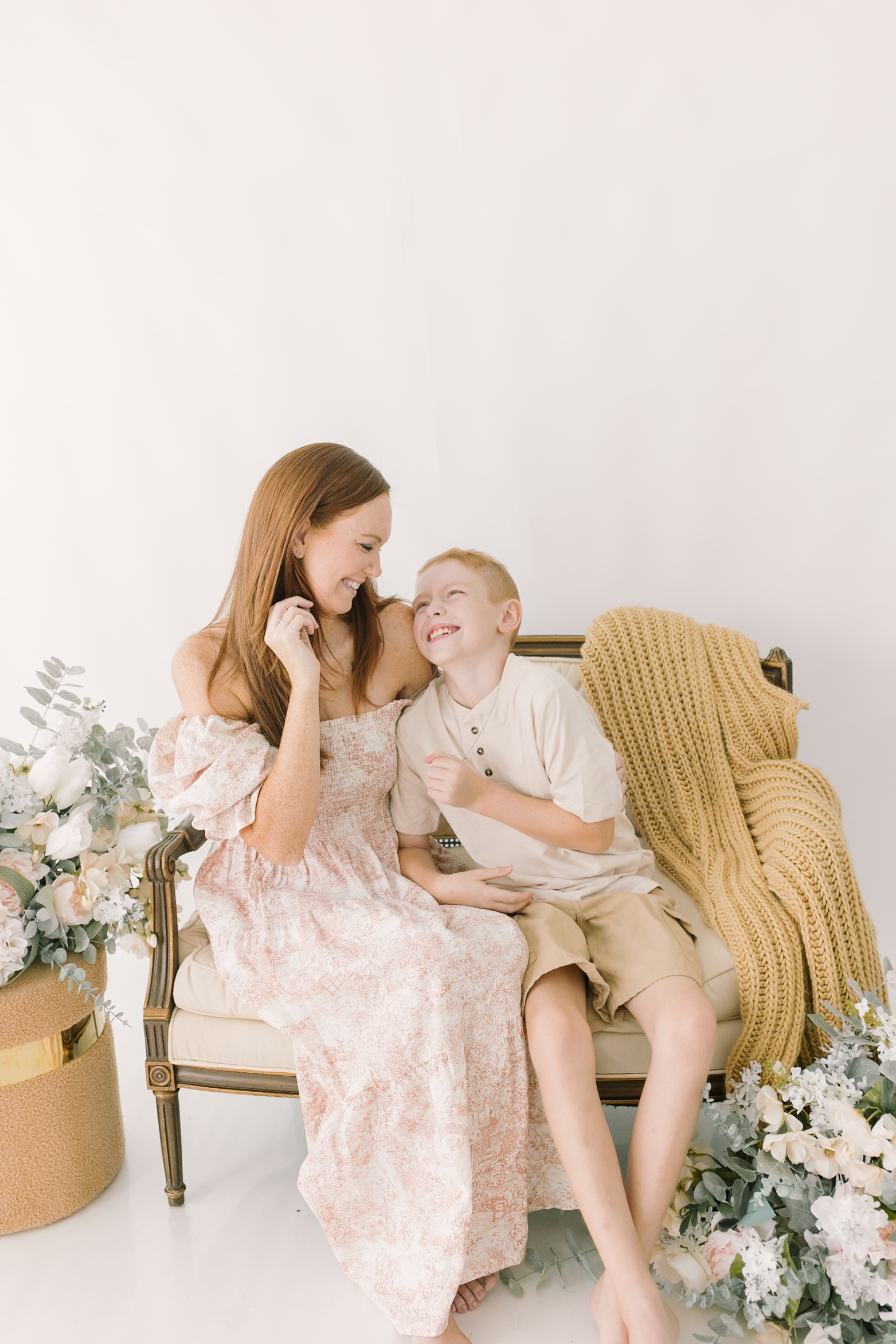 A happy mom in a floral print dress sits on a bench laughing with her toddler son in cream before visiting indoor playgrounds in raleigh