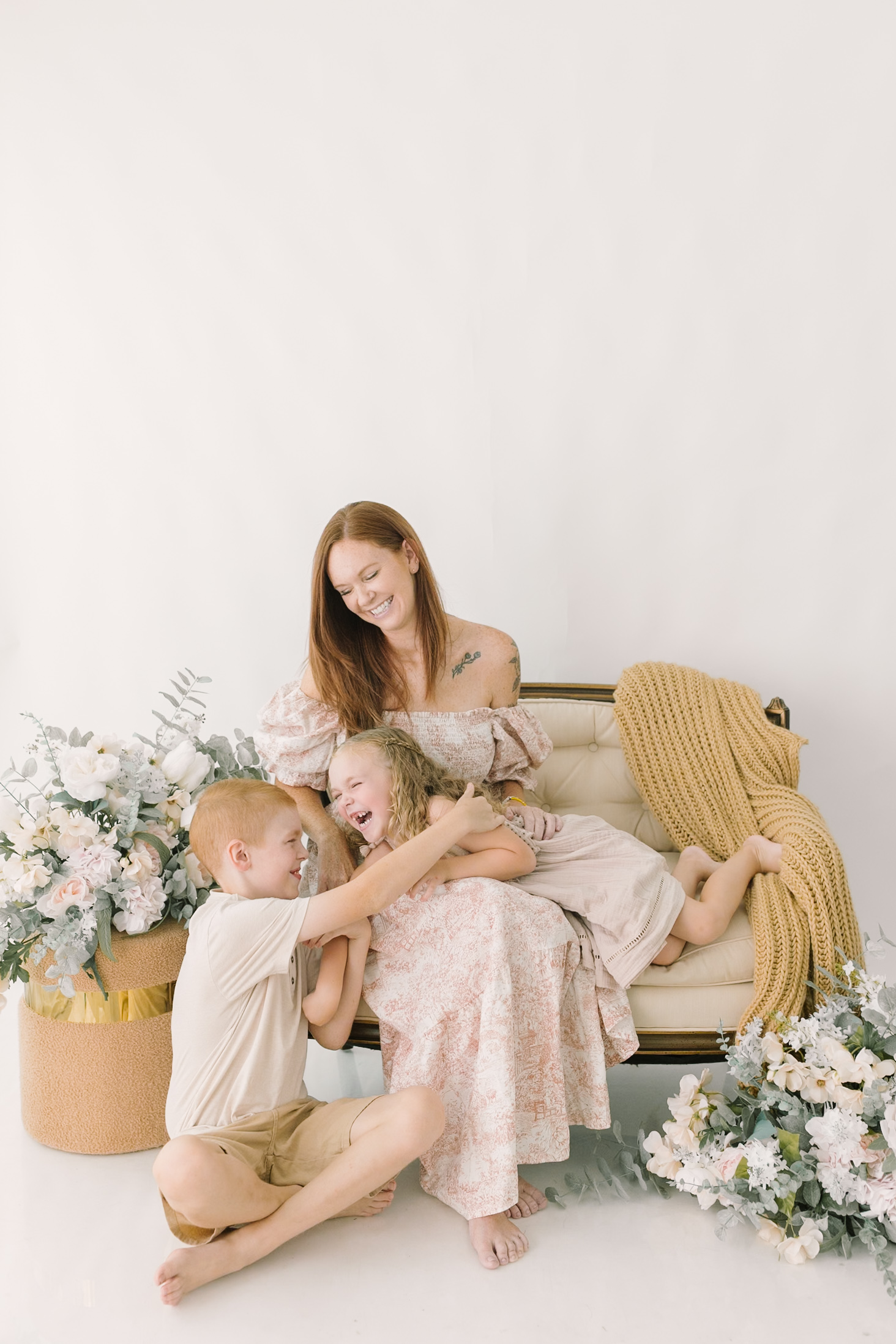 Toddler brother and sister play in mom's lap on a bench in a studio with white flowers before visiting a indoor playgrounds in raleigh