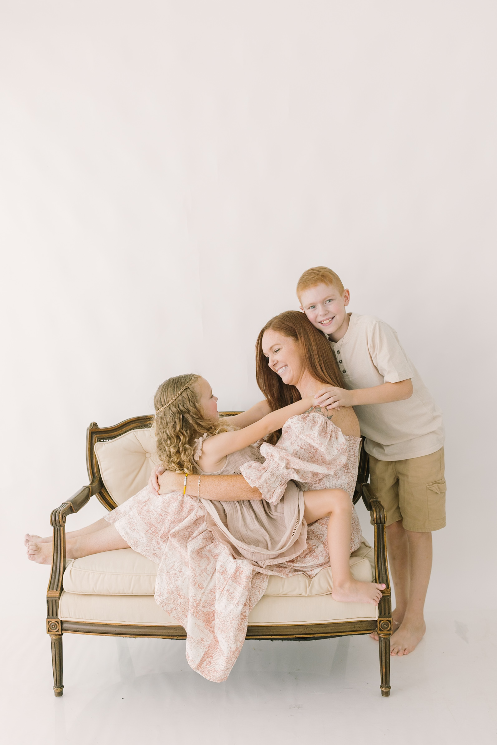 A happy mom sits across a vintage chair with her toddler daughter in her lap and young son hugging her while standing after visiting indoor playgrounds in raleigh