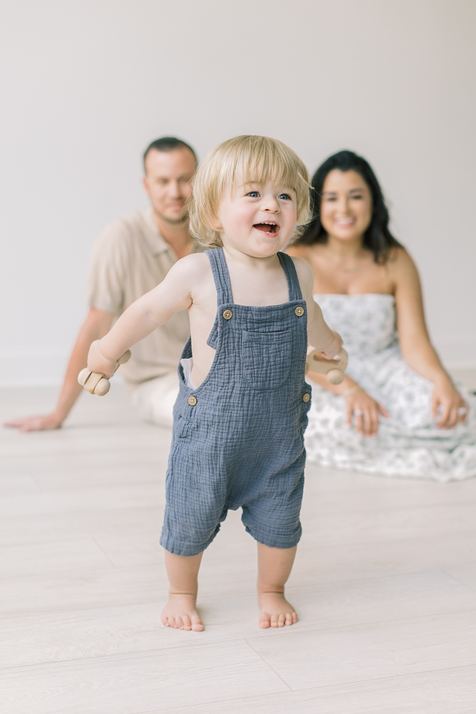 A happy toddler in blue overalls plays with wooden toys in a studio with mom and dad on the floor watching after visiting spots for a kid's birthday party in raleigh, NC