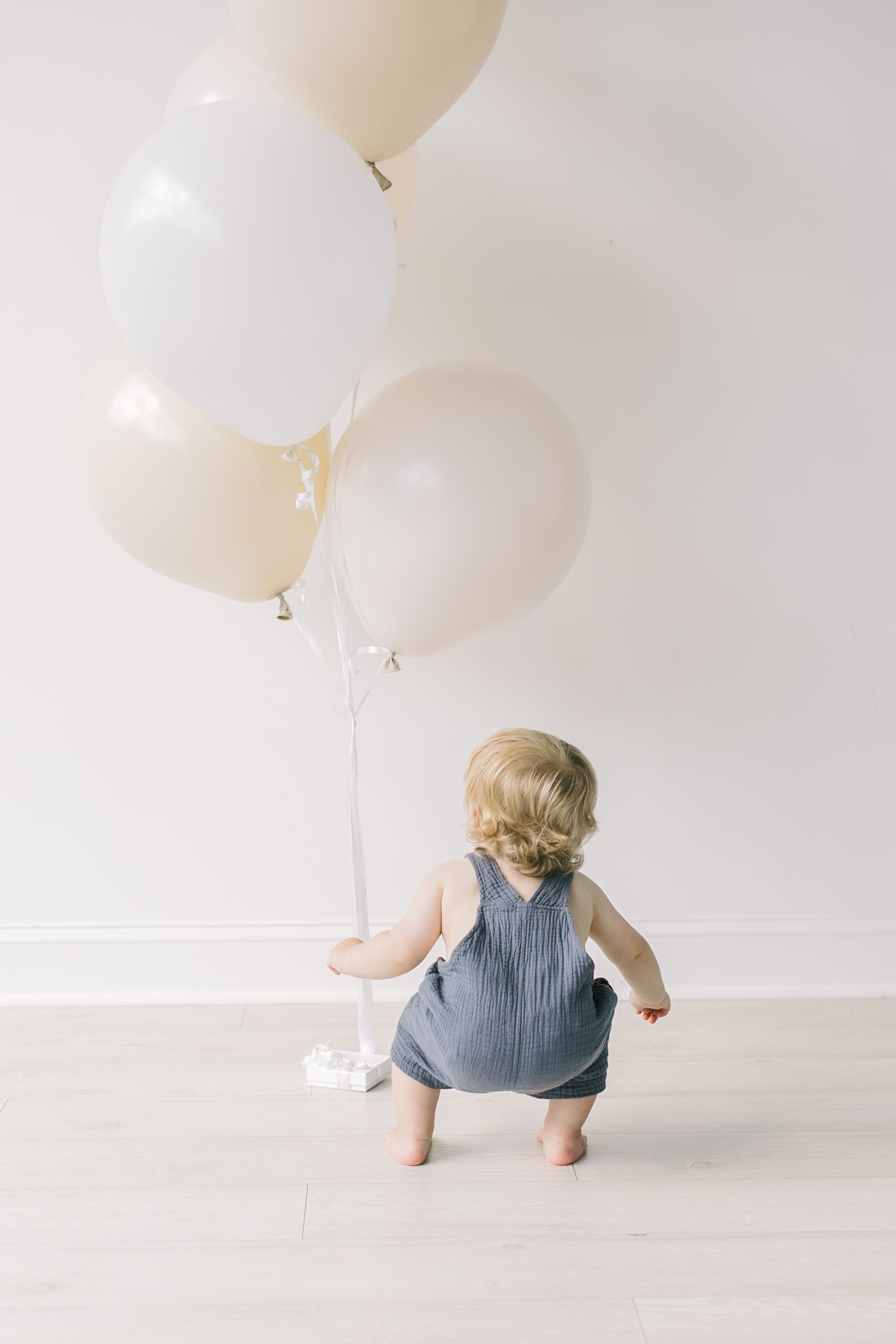 A happy toddler plays in blue overalls with some white balloons in a studio attached to a string